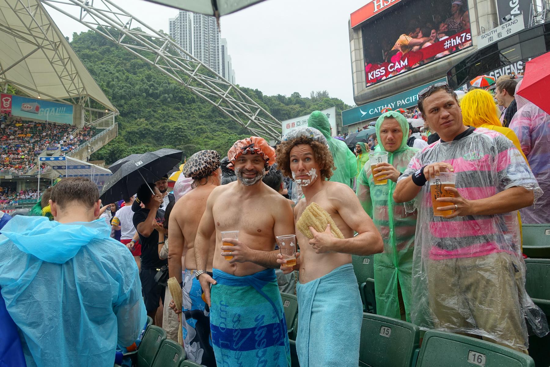 Rainy scene at the Hong Kong Rugby Sevens with spectators, including men in shower caps, towels, and shaving cream.