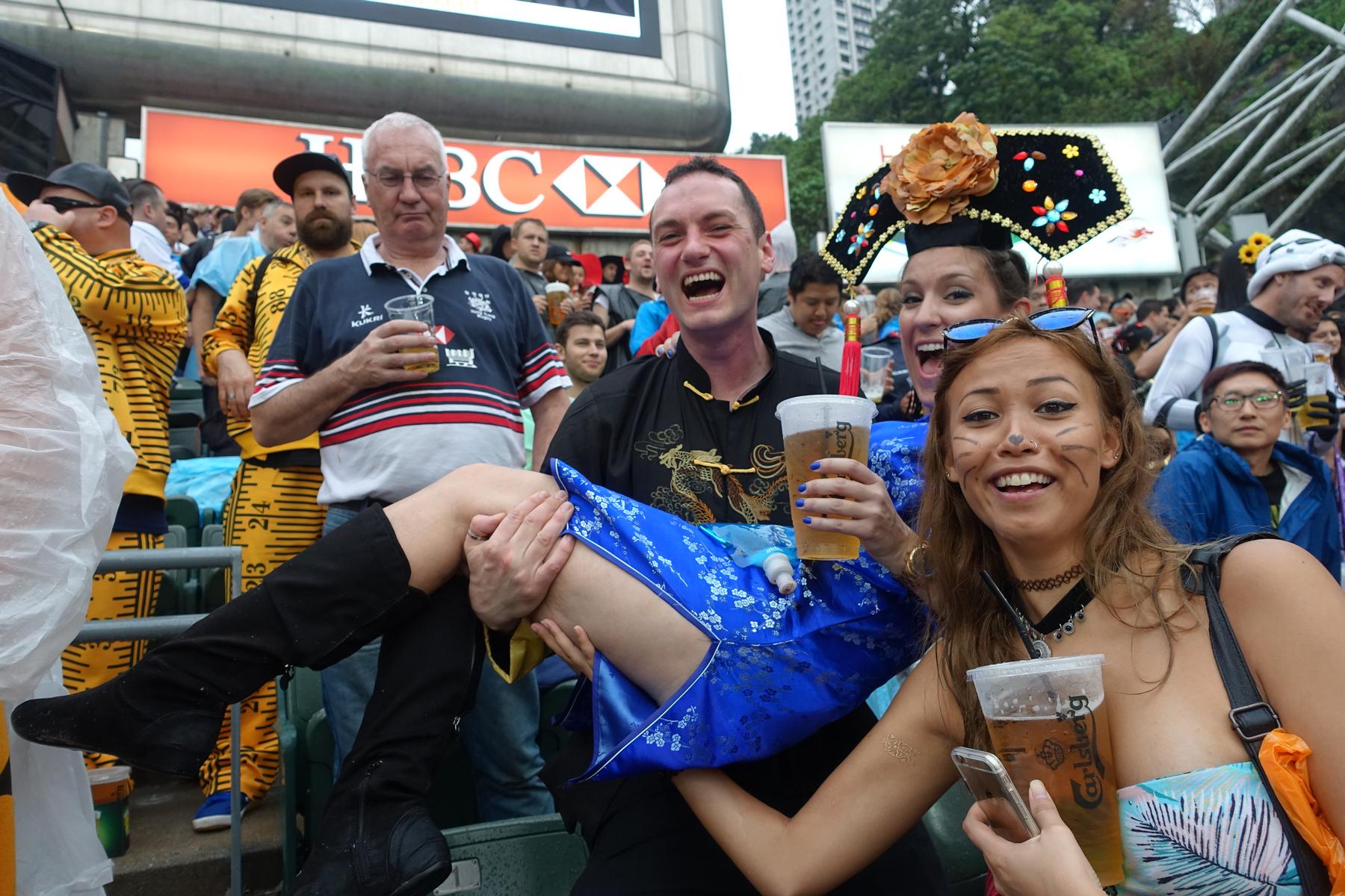 Jubilant rugby fans in costumes, including a man carrying a woman and another with face paint, hold beers at the Hong Kong Sevens.