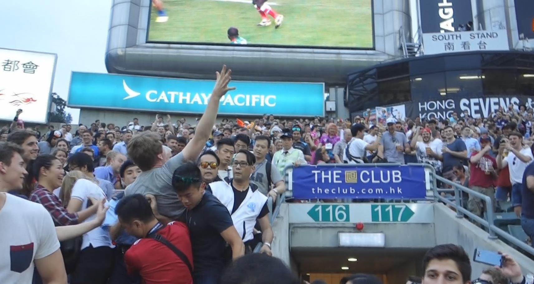 A dense crowd of people gathers in the South Stand area during the Hong Kong Rugby Sevens, with stadium signs and a game on a large screen.