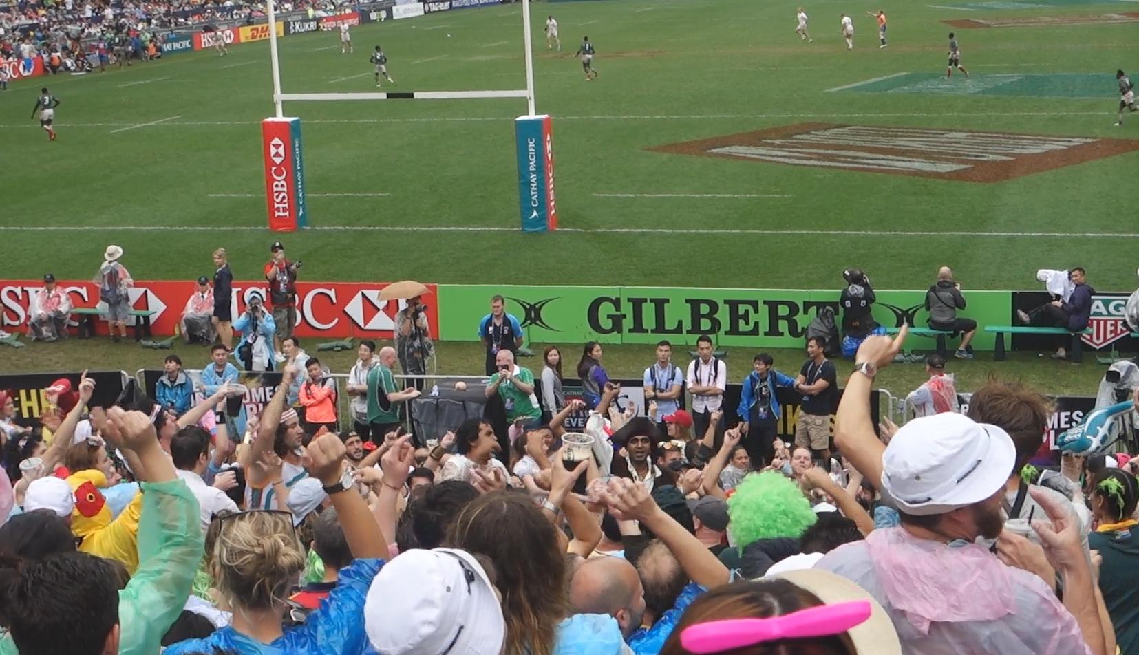 A large, lively crowd in the South Stand cheers at the Hong Kong Rugby Sevens match on a green field.
