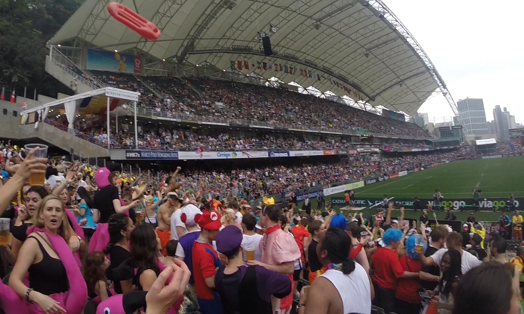 A festive, costumed crowd cheers enthusiastically in a packed stadium during the Hong Kong Rugby Sevens.