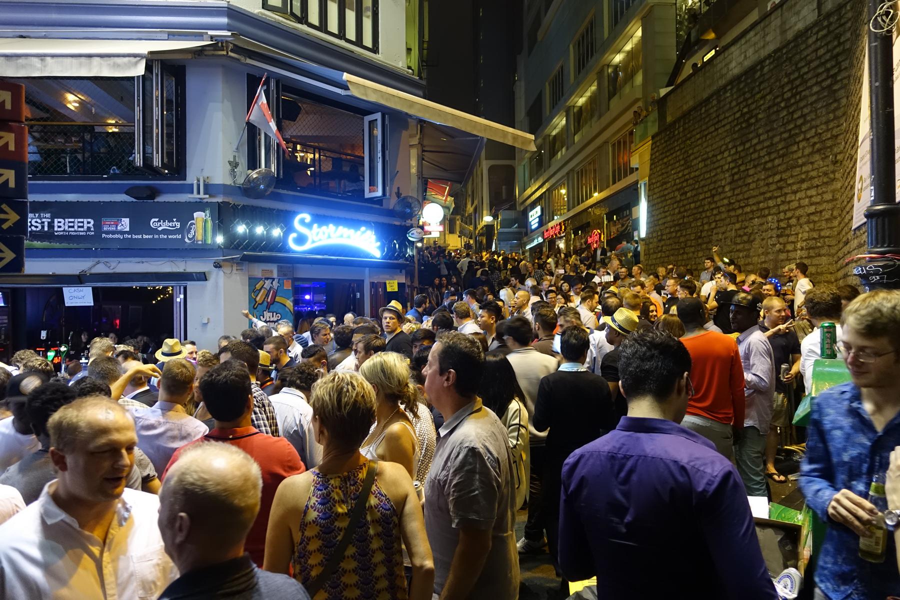 A lively street scene at night with a large crowd gathered outside illuminated bars.