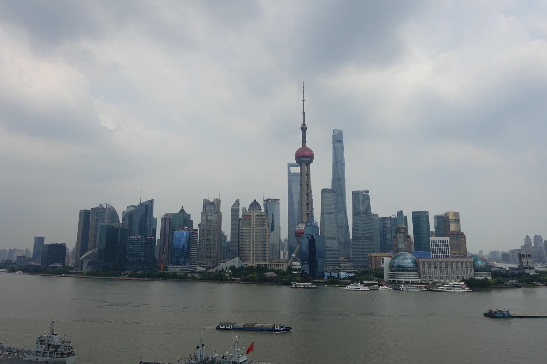 A panoramic view of the Shanghai skyline, including the Oriental Pearl TV Tower, across the Huangpu River with boats.