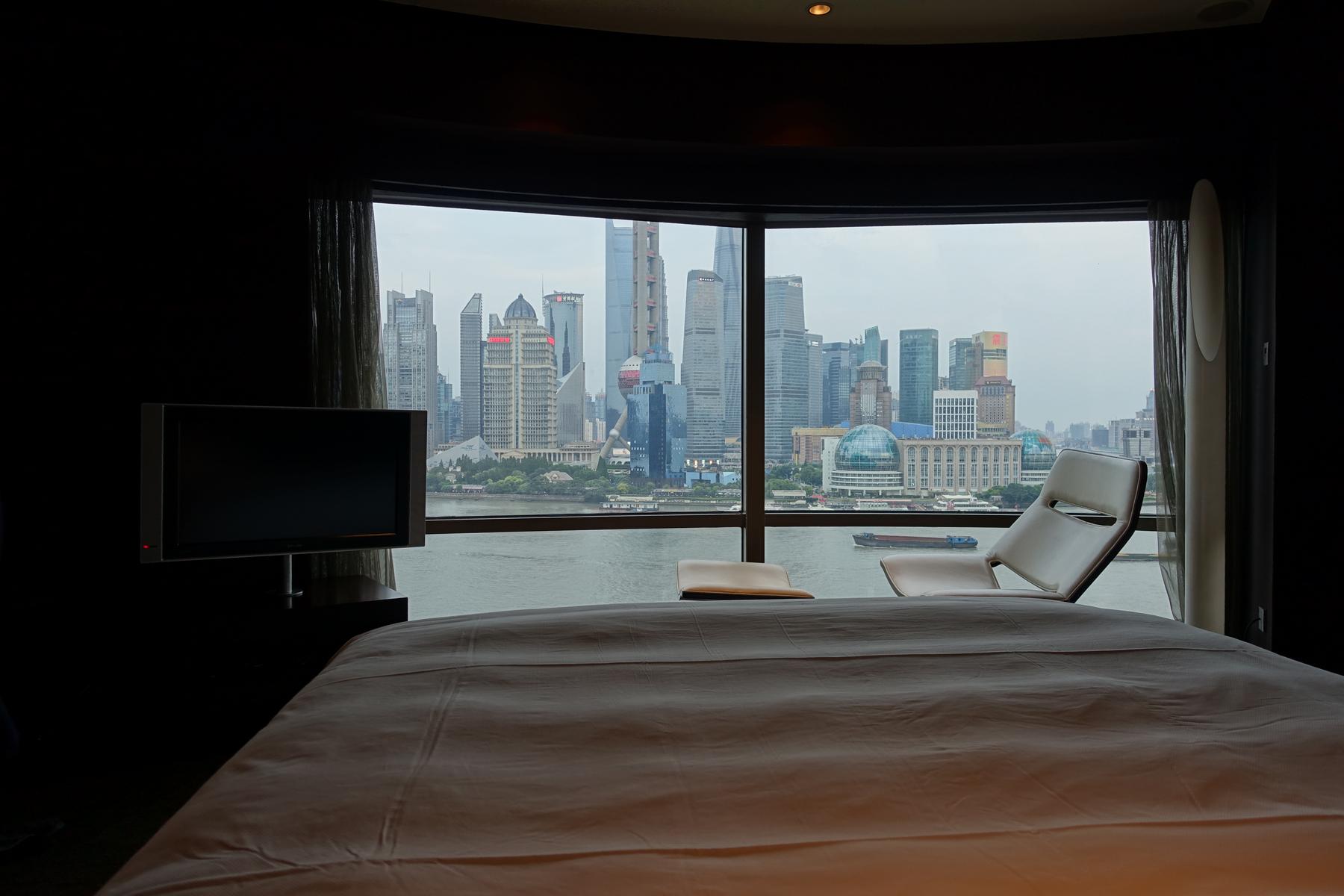 Panoramic view of the Shanghai skyline across the Huangpu River from a hotel room with a bed and TV.