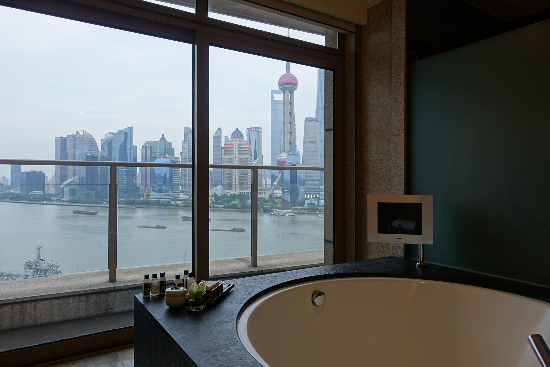A hotel bathtub with a panoramic view of the Shanghai skyline and Huangpu River through a large window.