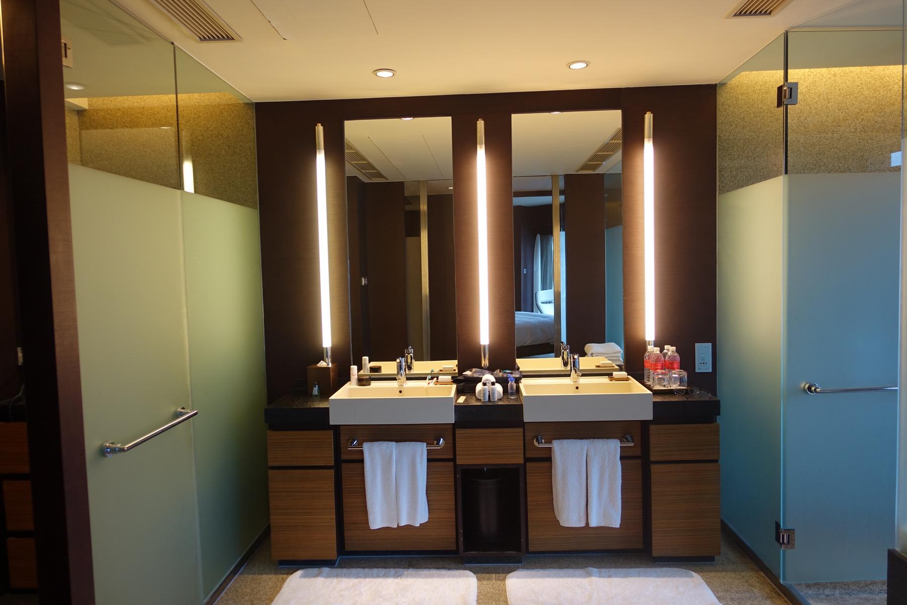 A modern hotel bathroom features a dark wood double vanity with two sinks, bright vertical lights, large mirrors, and frosted glass walls.