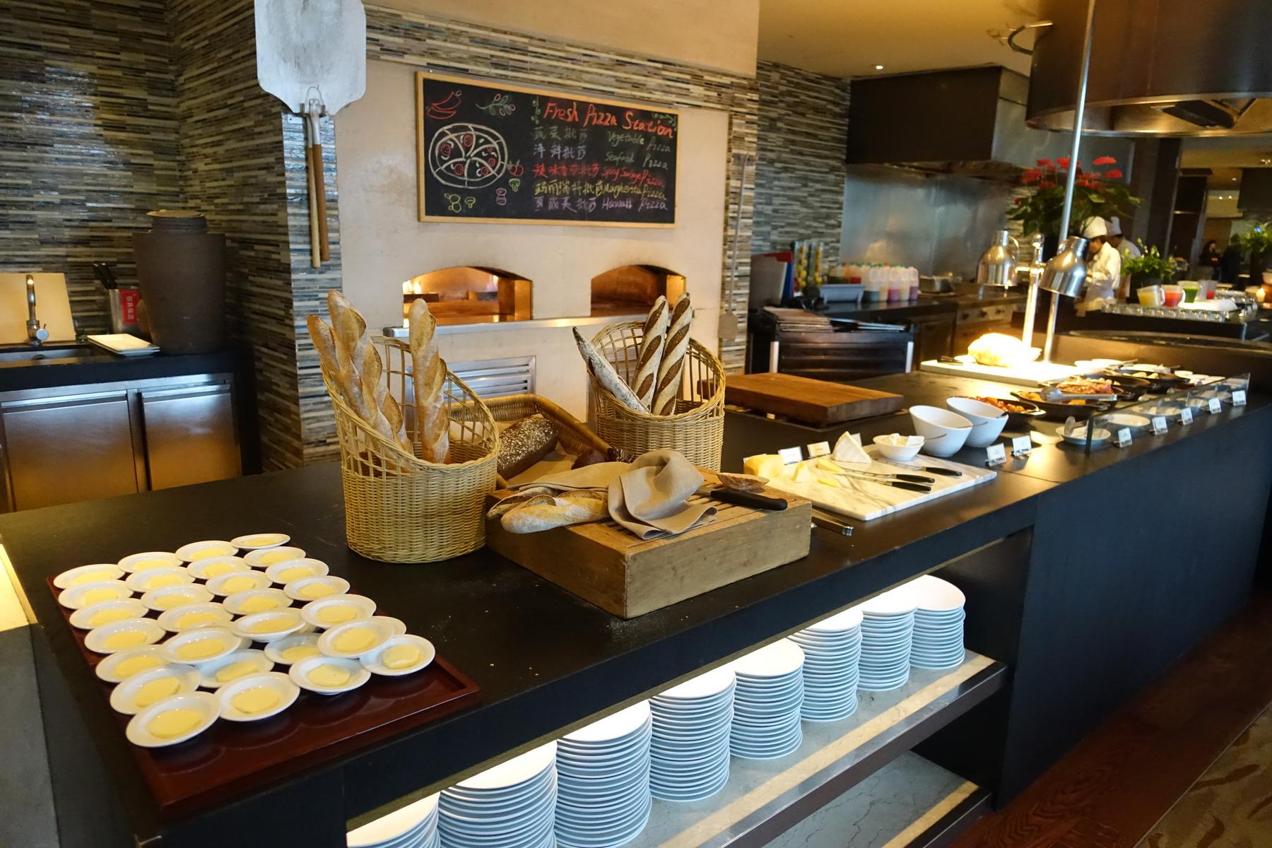 A hotel buffet featuring a "Fresh Pizza Station" with wood-fired ovens, baskets of bread, cheeses, and various prepared dishes.