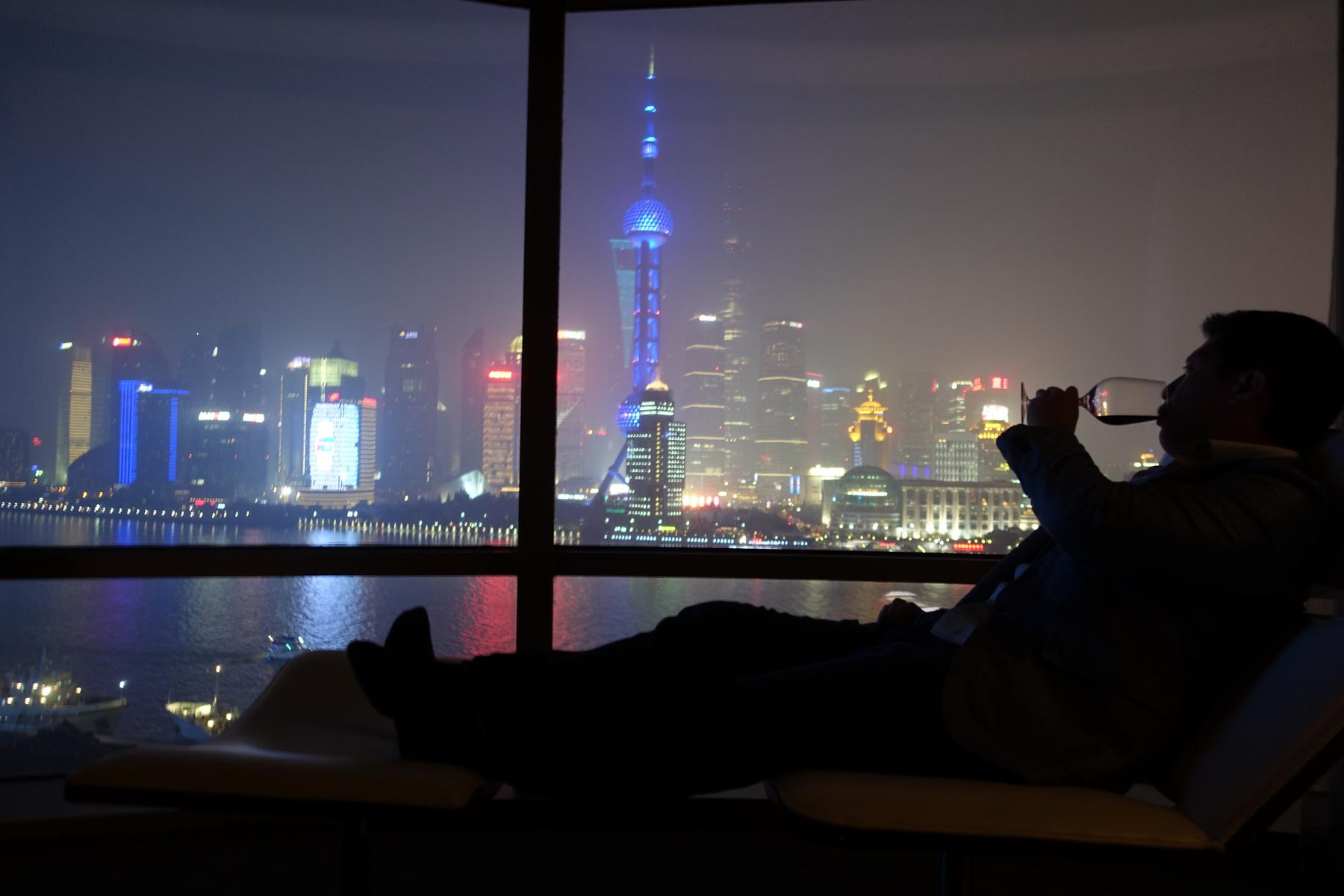 A person sips wine from a glass while relaxing and looking out at the illuminated Shanghai skyline at night, featuring the Oriental Pearl TV Tower.