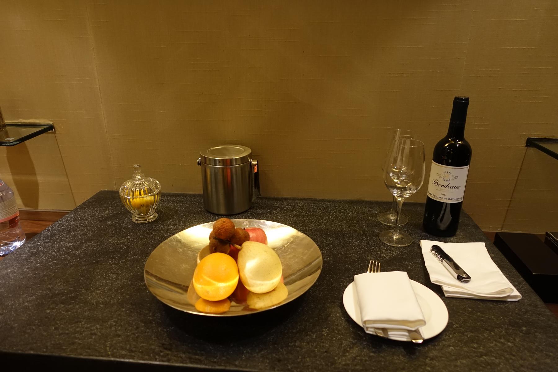 A hotel welcome amenity featuring a fruit platter, a bottle of red wine with glasses, and table settings on a dark table.