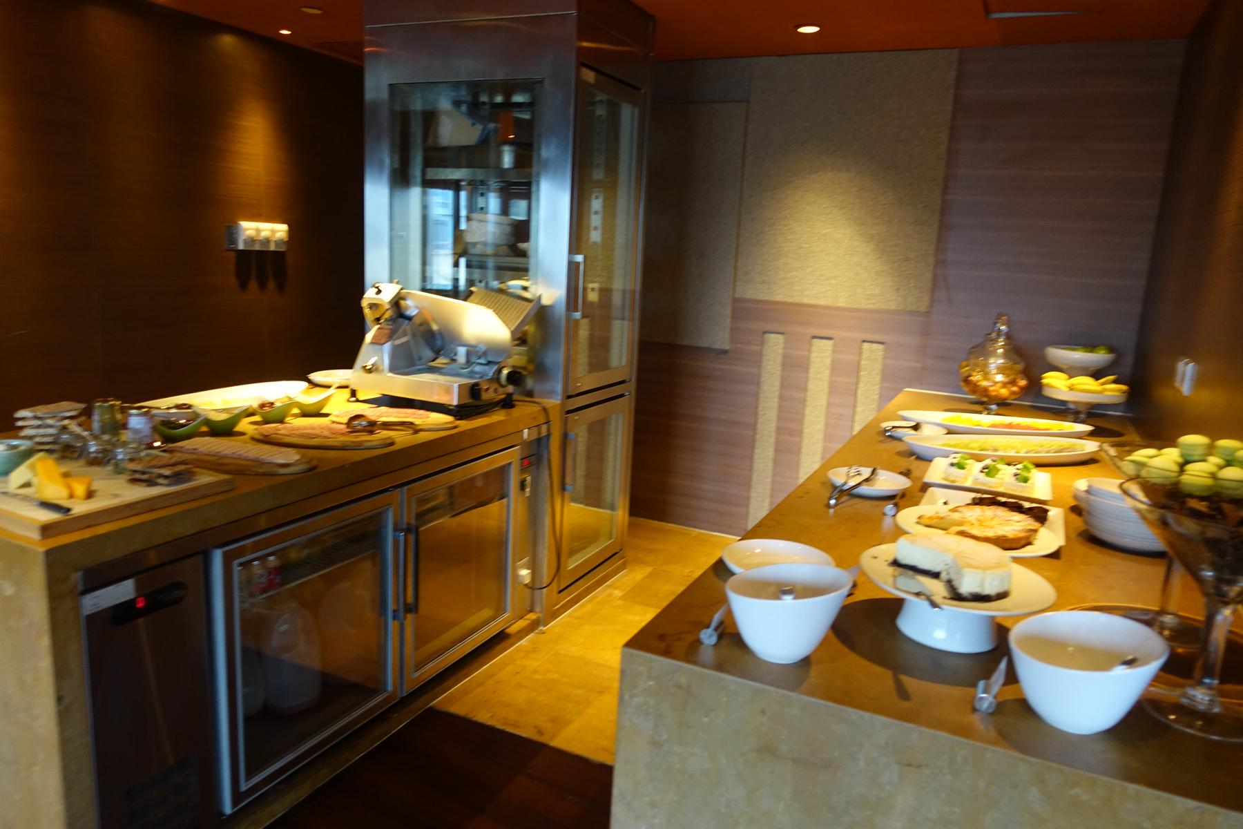 A hotel buffet spread featuring a meat slicer, cheeses, fruits, and an assortment of desserts including cakes and macarons.