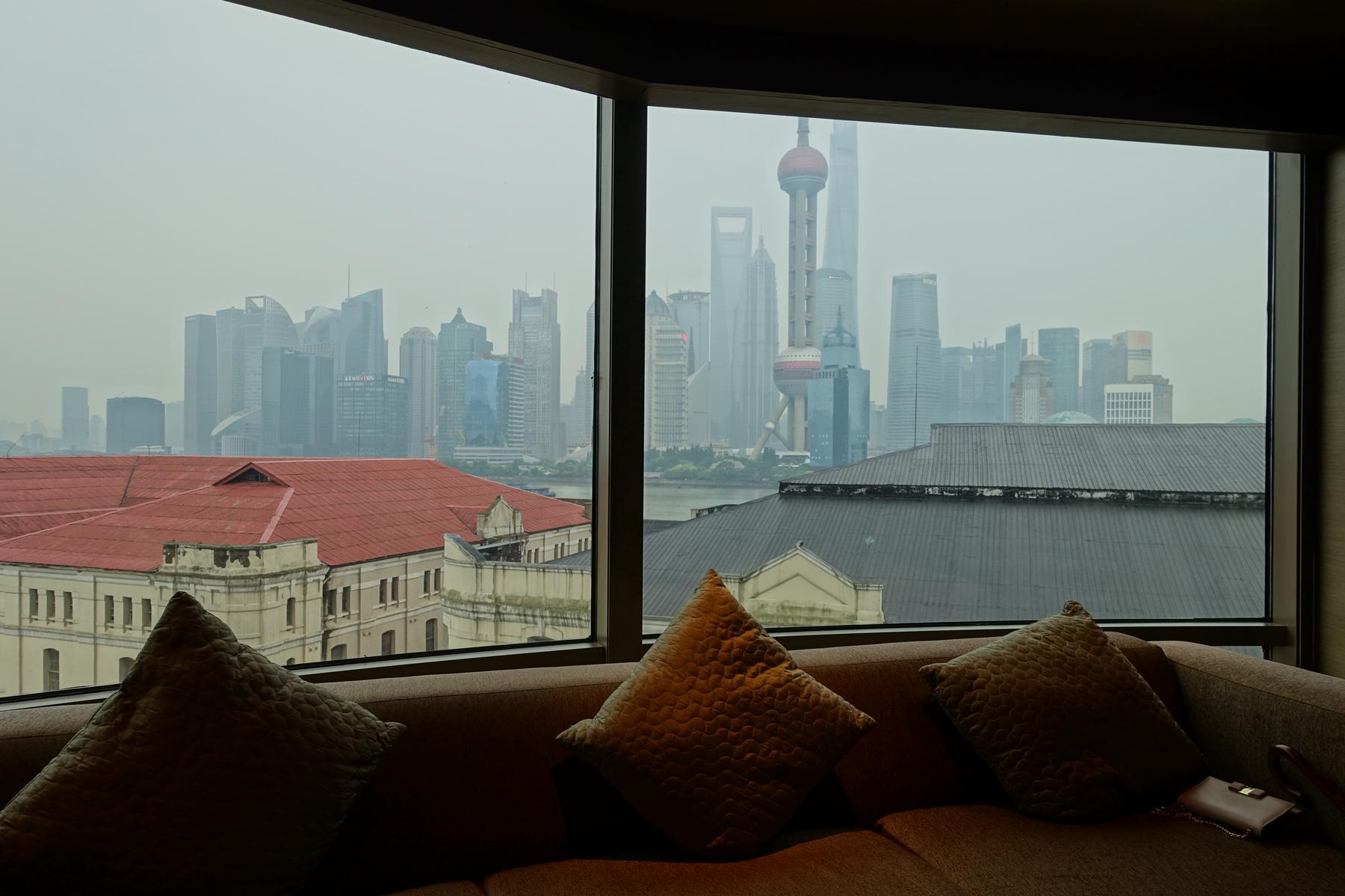 A hotel room couch with pillows looks out to a hazy Shanghai skyline and the Oriental Pearl TV Tower.