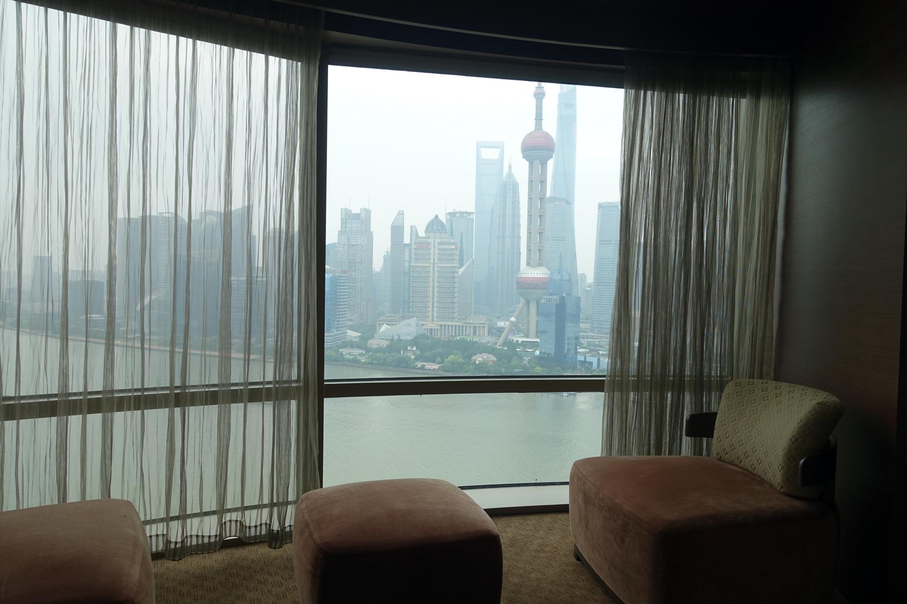 A hotel room window frames a view of the Shanghai skyline, including the Oriental Pearl TV Tower and the Huangpu River, with two ottomans in the foreground.