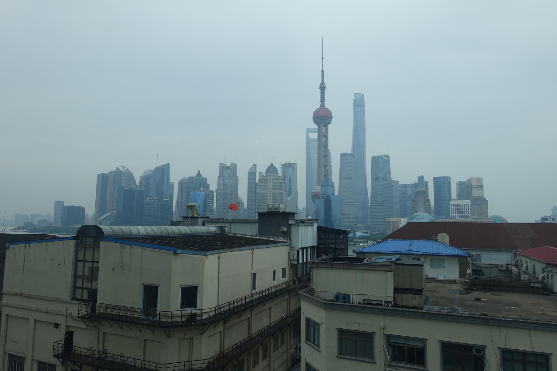 Hazy view of the Shanghai skyline with the Oriental Pearl TV Tower and skyscrapers rising above foreground buildings.