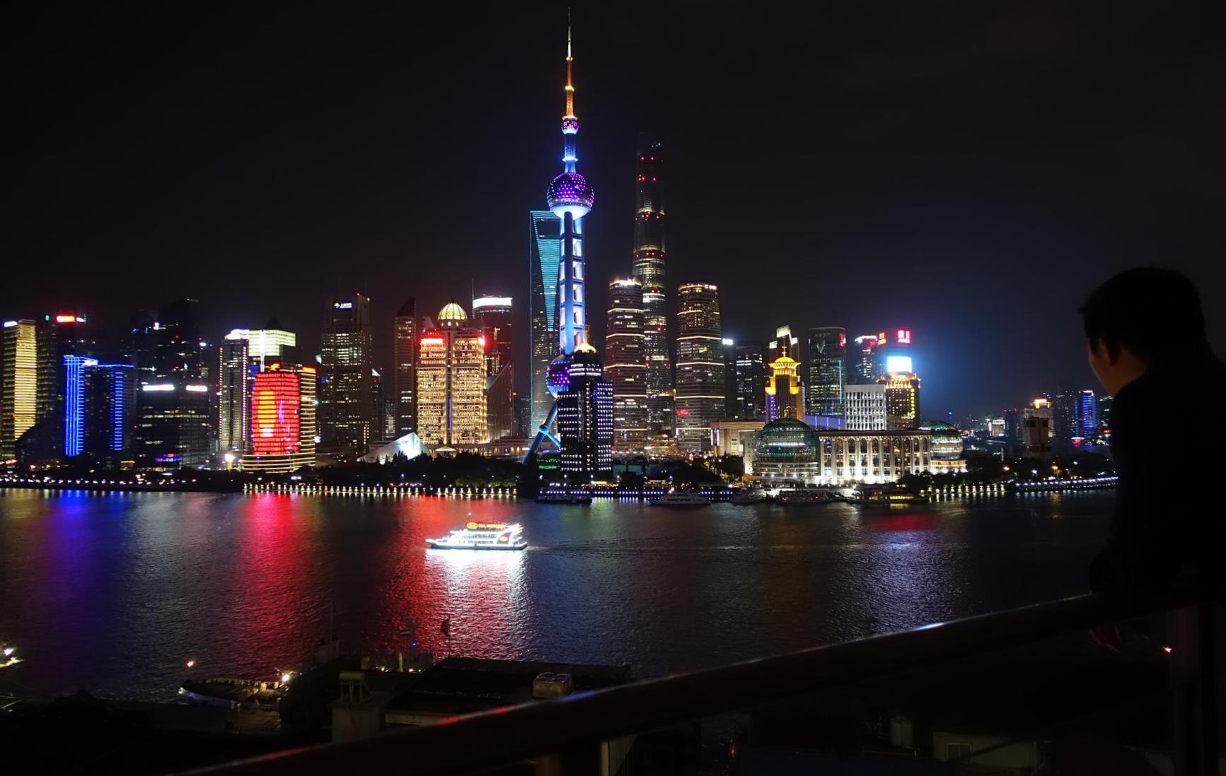 Night view of the brightly lit Shanghai Bund skyline and Huangpu River with a person looking out.