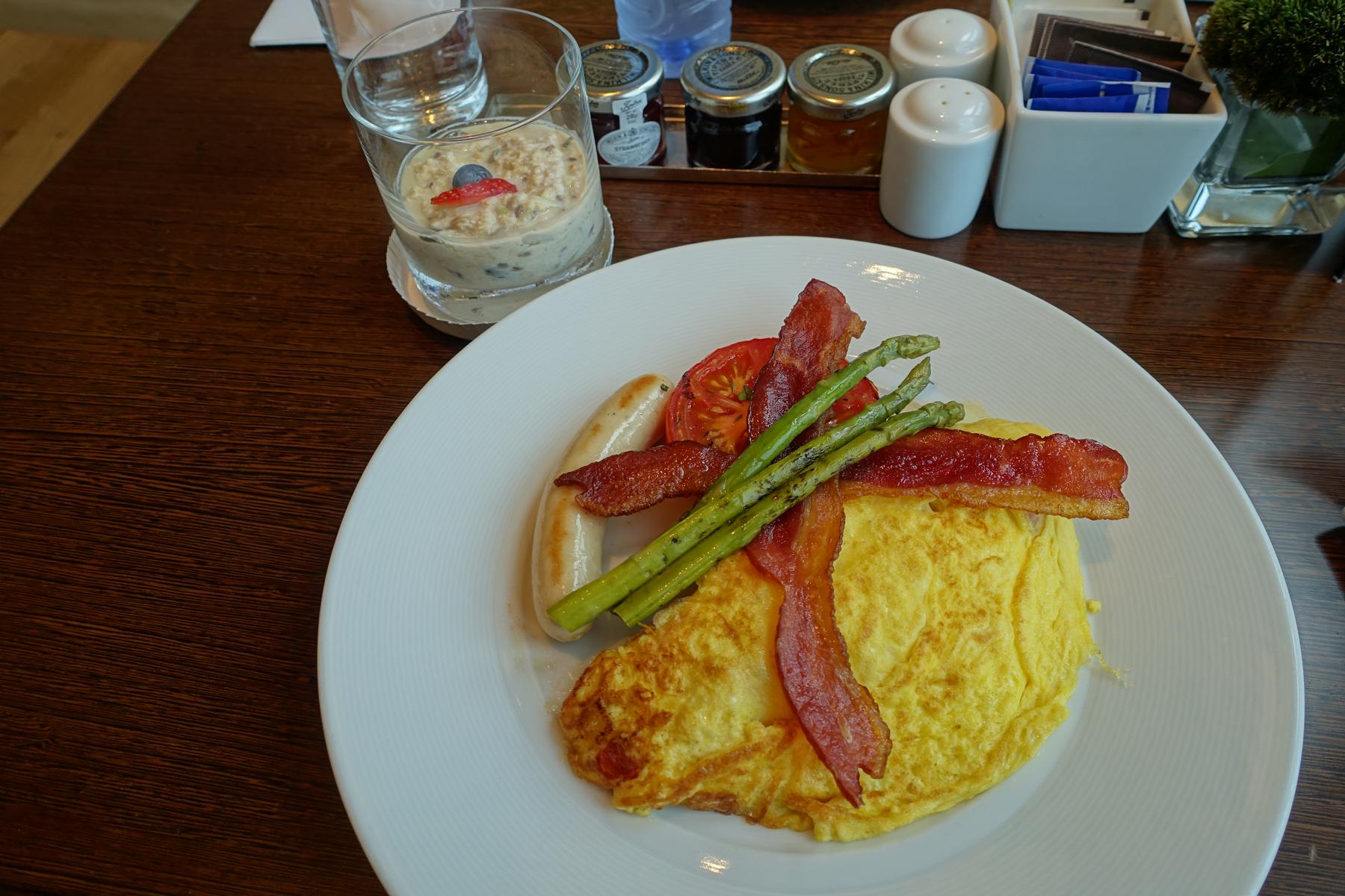 A breakfast plate with an omelet, bacon, sausage, asparagus, and grilled tomato, alongside a glass of overnight oats.