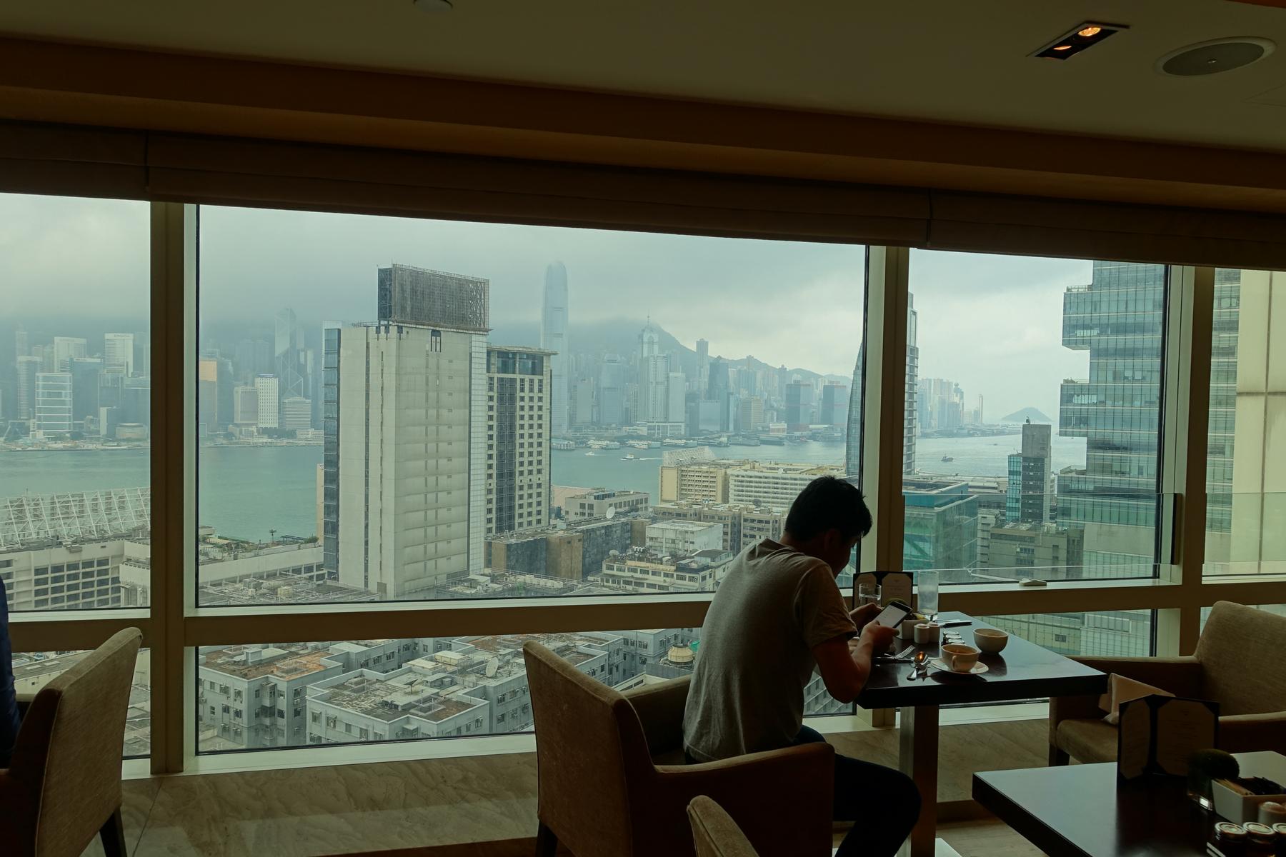 A person sits at a table by a window, looking at a phone, with an overcast view of the Hong Kong skyline and harbor.