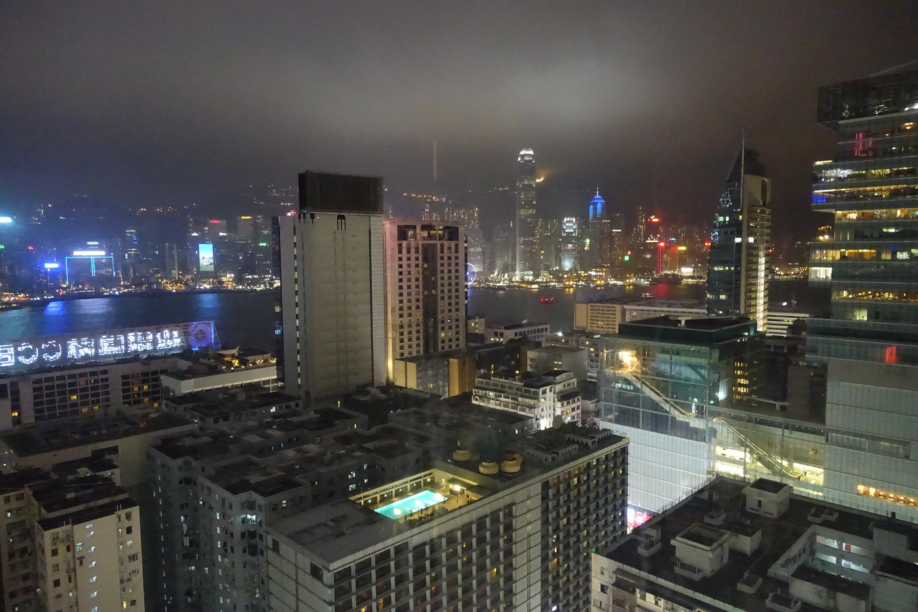 Nighttime cityscape of Hong Kong from above, featuring Victoria Harbour, brightly lit skyscrapers, and a rooftop pool.