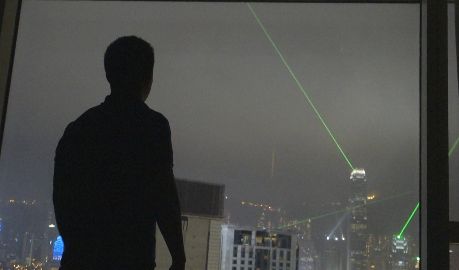 A silhouetted person looks out a window at the Hong Kong skyline with green laser beams at night.