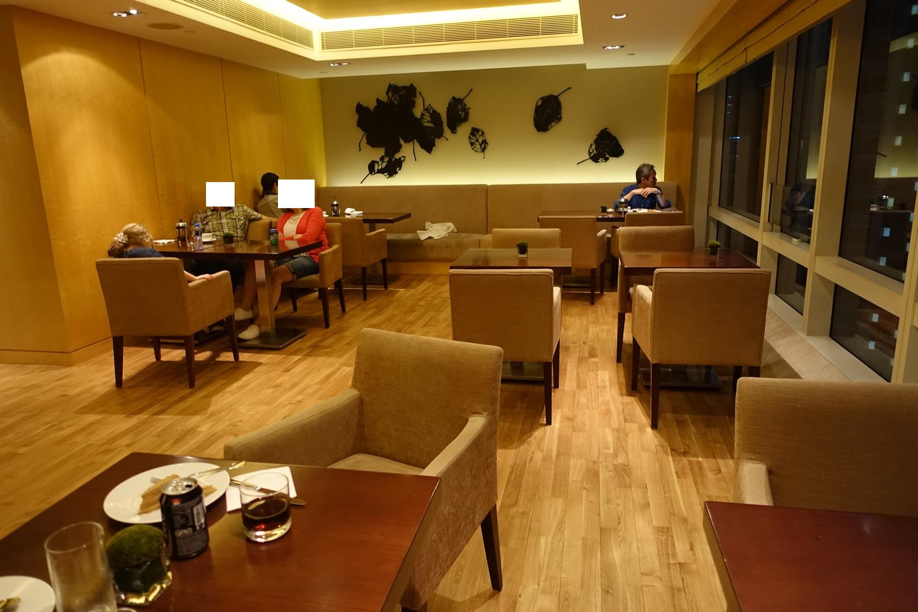 A hotel lounge or restaurant with guests dining at tables and a city night view through large windows.