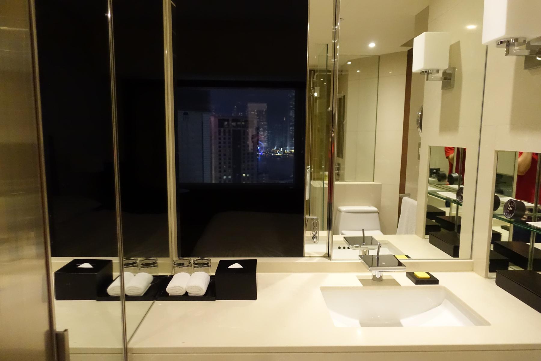 Hotel bathroom vanity with a mirror reflecting a city night view and bedroom.