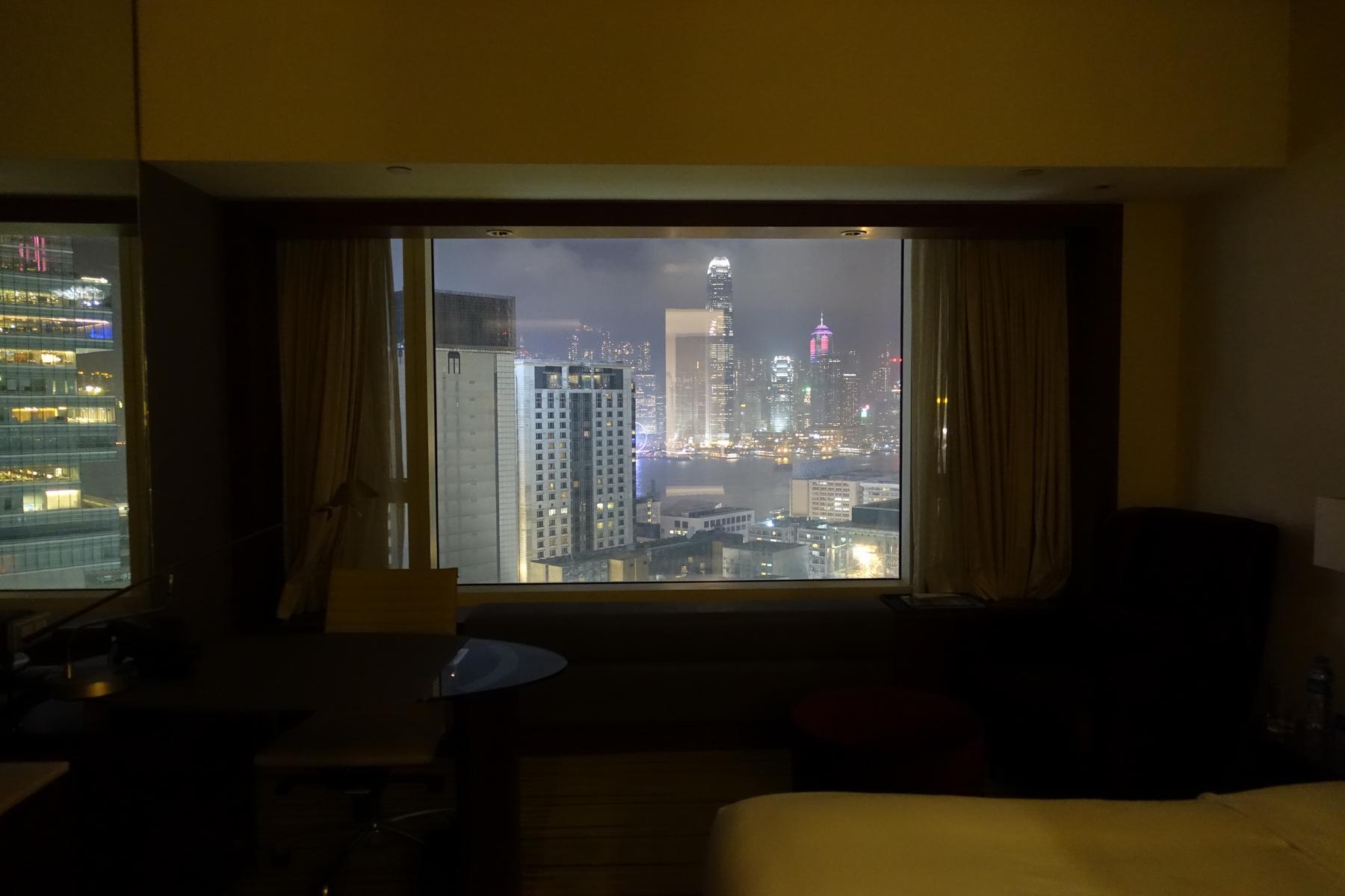 Night view from a hotel room showing the illuminated Hong Kong cityscape and harbor through a large window.