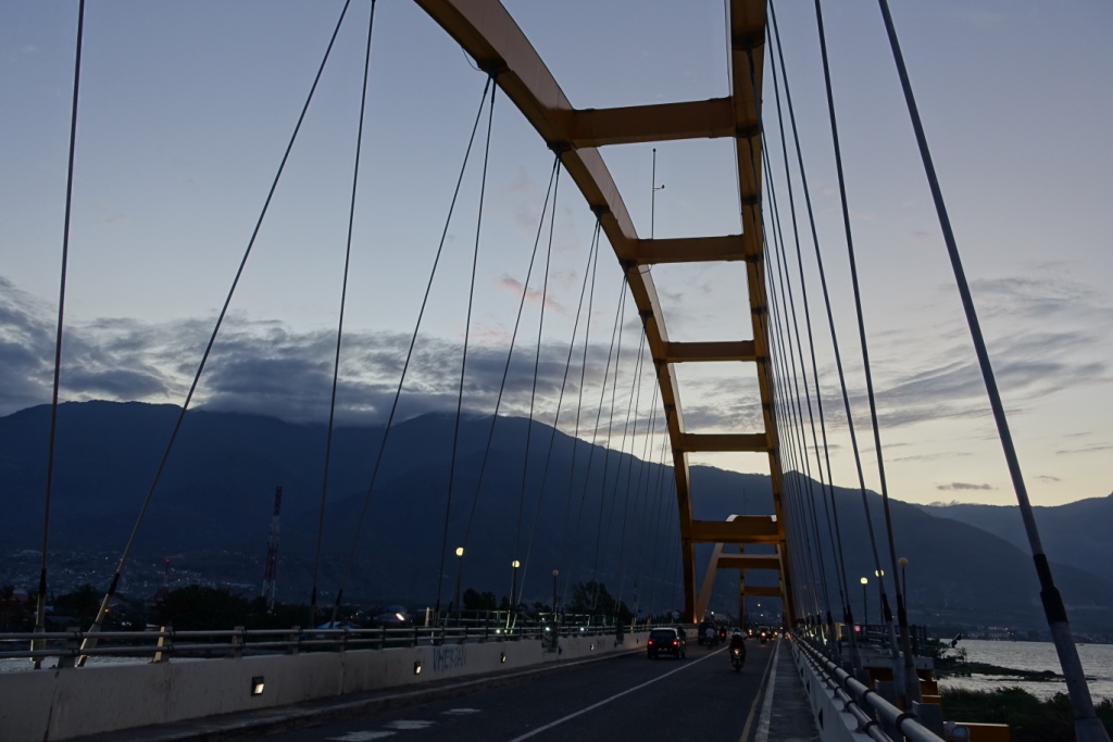 A yellow arch bridge carries a road with vehicles over water, with dark mountains under a twilight sky.