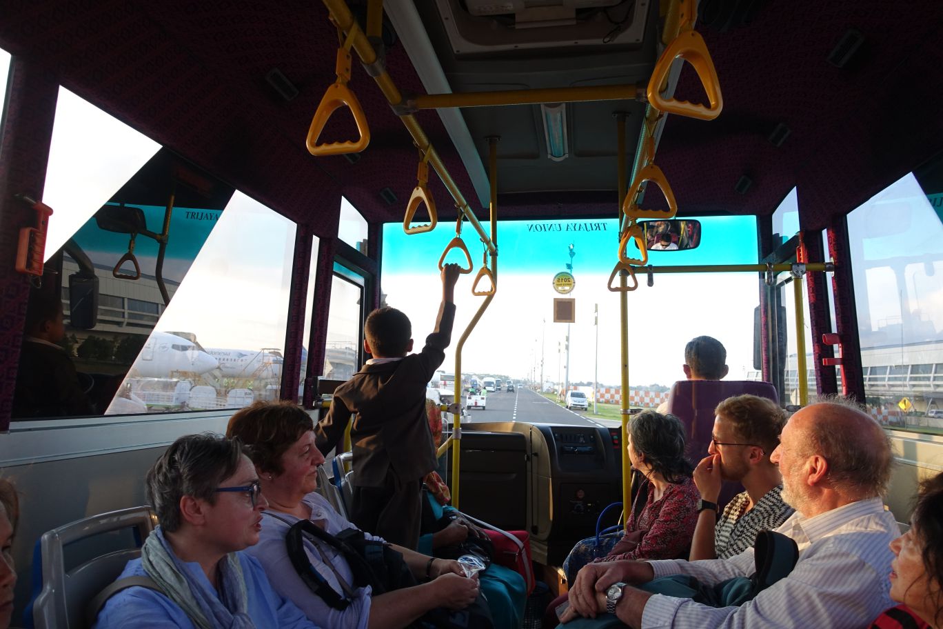 Inside an airport shuttle bus, passengers look out as an airplane and airport road are visible through the windows.