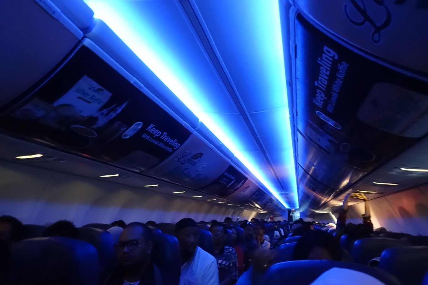 Inside a Lion Air plane with blue mood lighting, showing overhead advertisements and seated passengers.