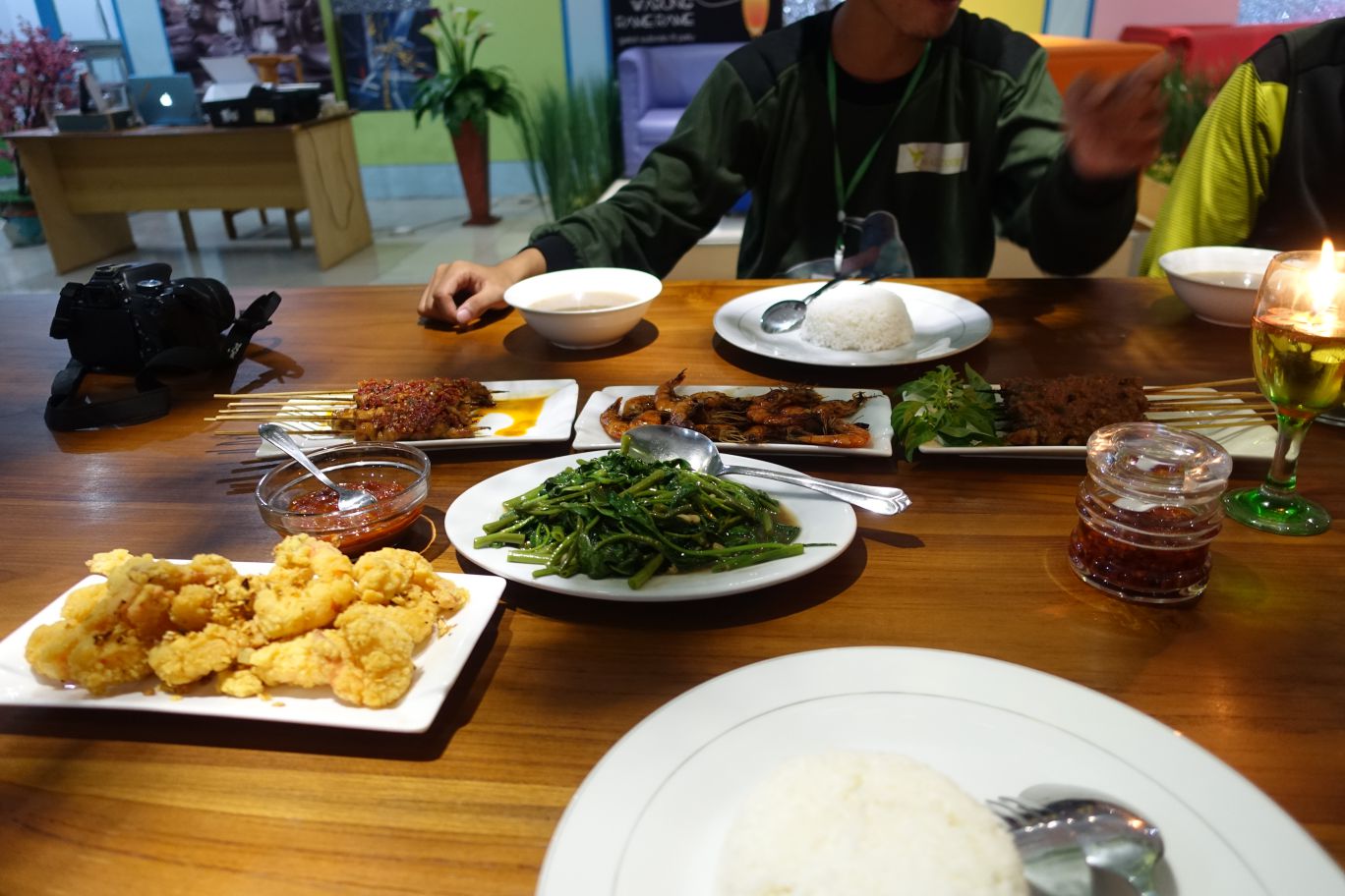 A dining table laden with various Indonesian dishes including fried calamari, satay, greens, and rice, with a person's hands visible.