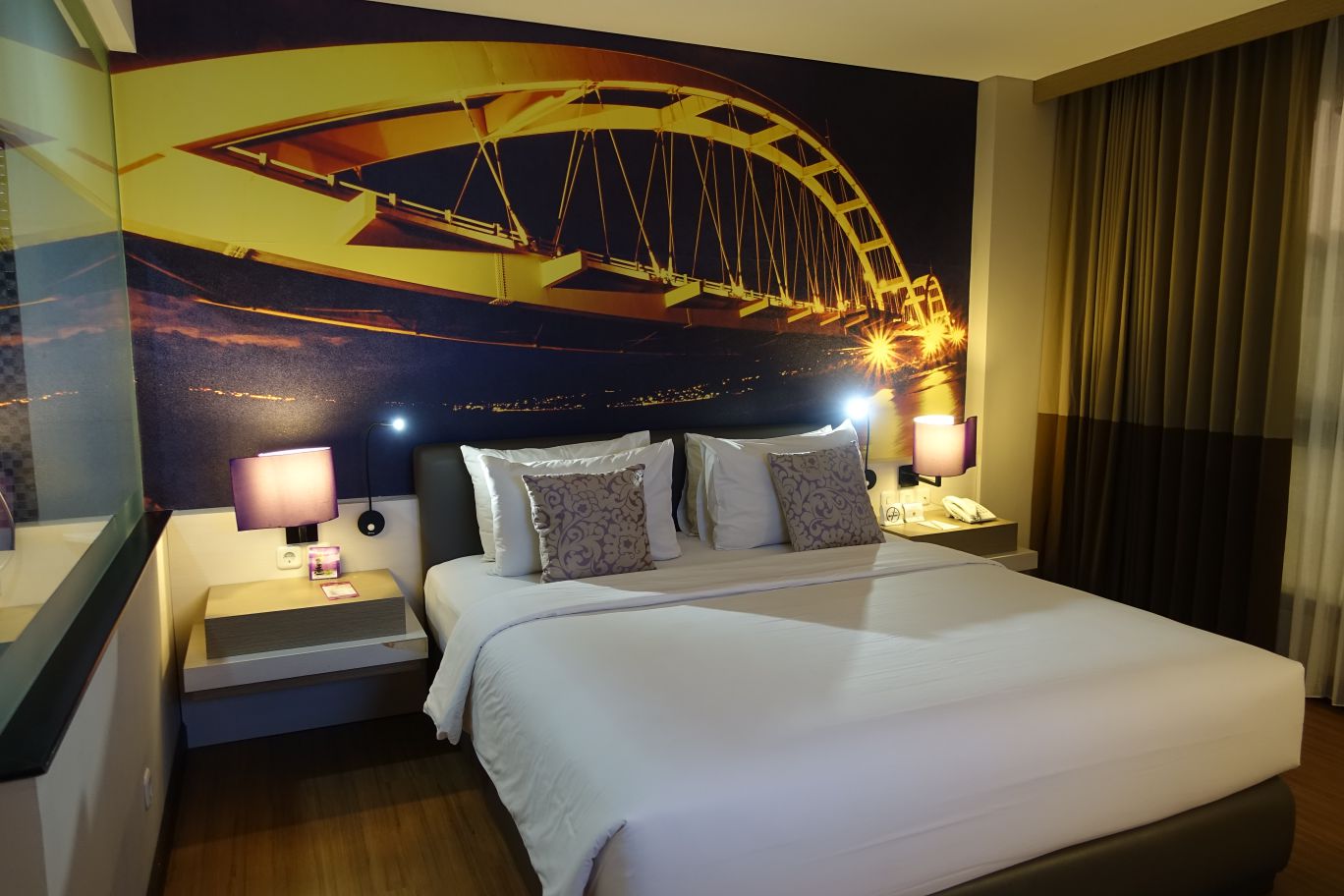 A modern hotel room with a large bed and a wall mural of an illuminated bridge at night.