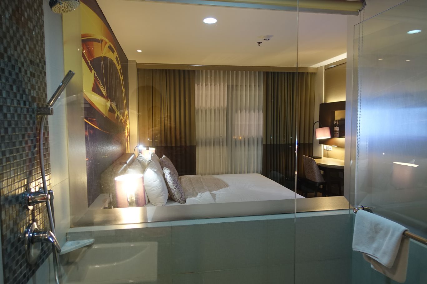 A modern hotel bedroom with a bed, desk, and large window is viewed through a glass partition from its adjacent bathroom with a shower.