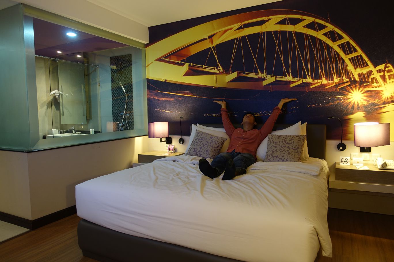 A man lies on a hotel bed with his arms raised, a large mural of a lighted bridge behind him, and a glass-walled bathroom visible to his left.