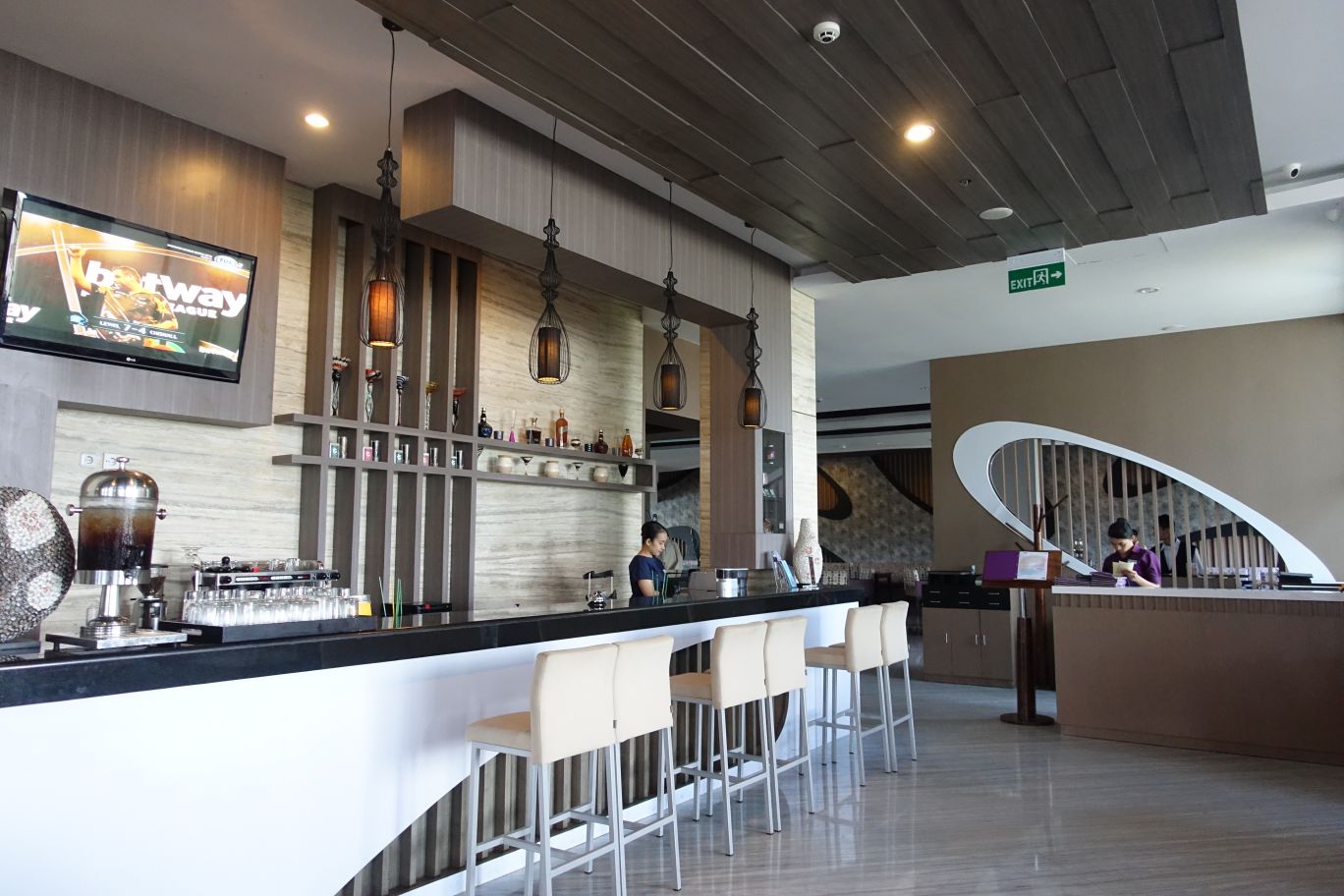 A modern hotel bar area with a long counter, bar stools, a TV, and a reception desk in the background.