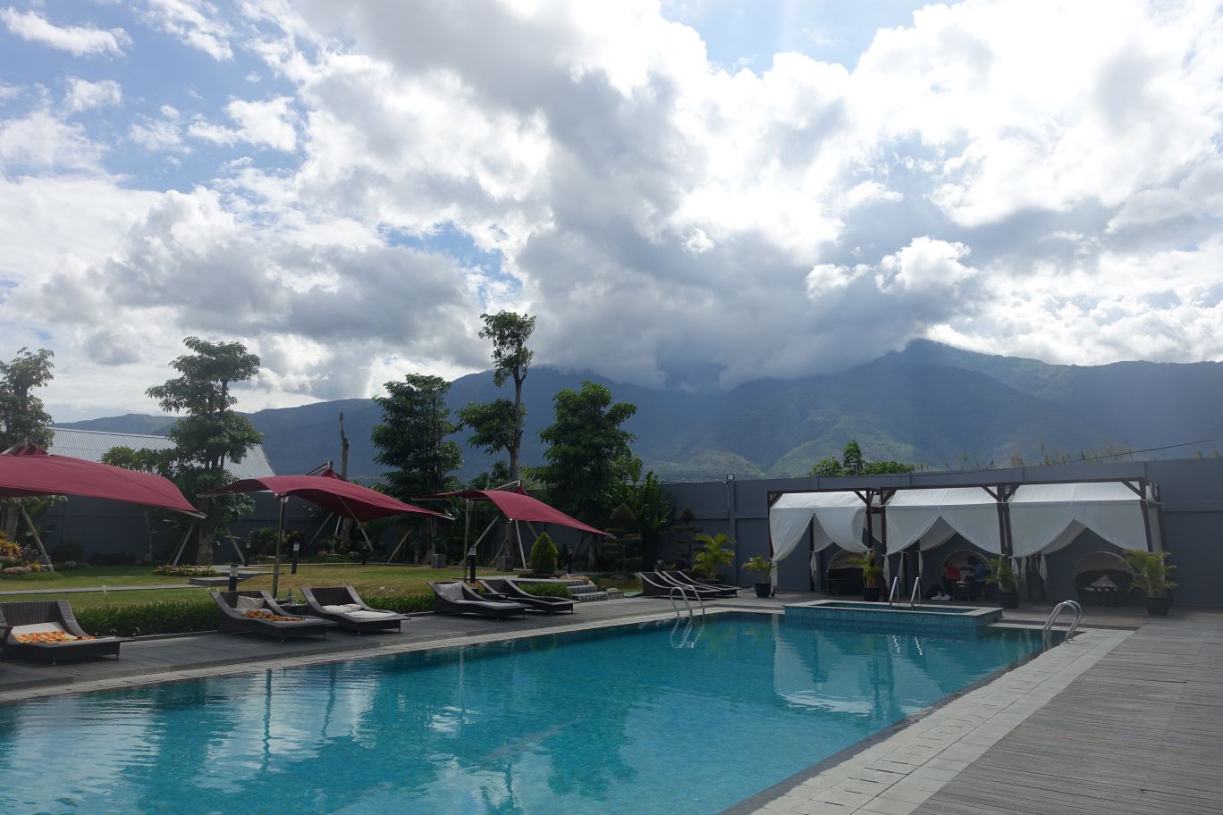 An outdoor swimming pool with lounge chairs, red umbrellas, and white cabanas, backed by mountains and a cloudy sky.