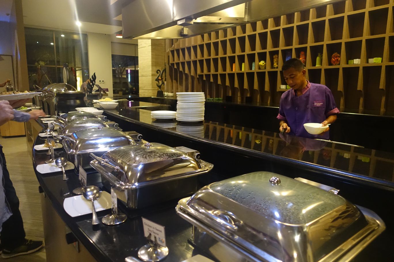 A chef stands behind a buffet line with multiple chafing dishes in a hotel restaurant.