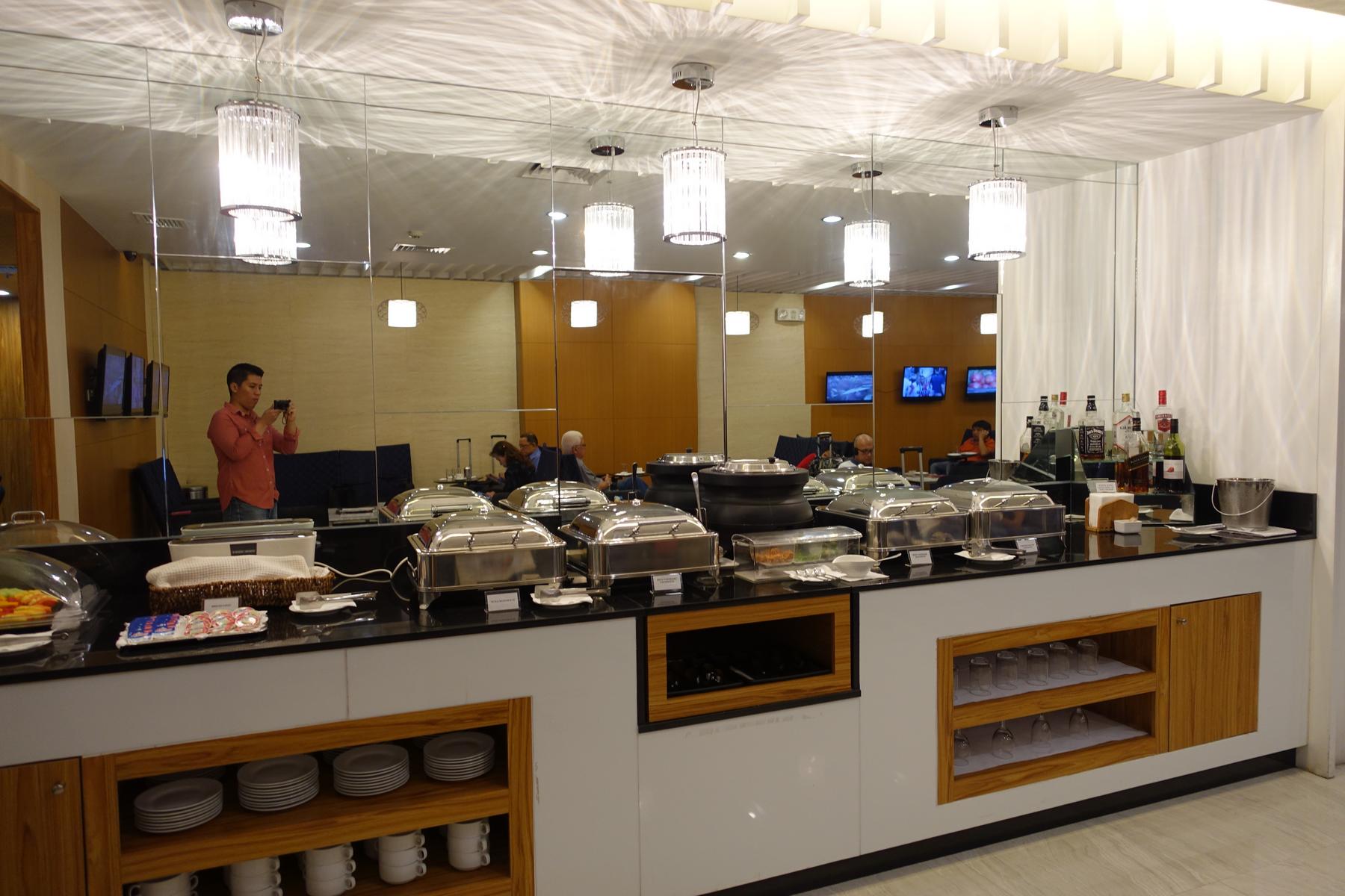 Chafing dishes and drinks on a buffet counter in a mirrored airport lounge.