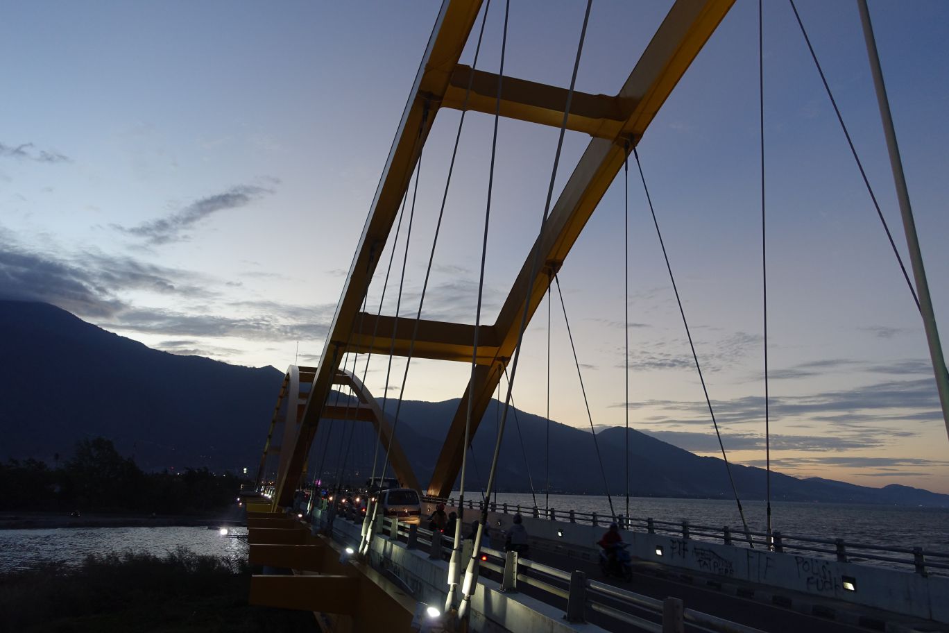 Ponulele Bridge in Palu, Indonesia, at dusk with traffic, water, and mountains in the background.