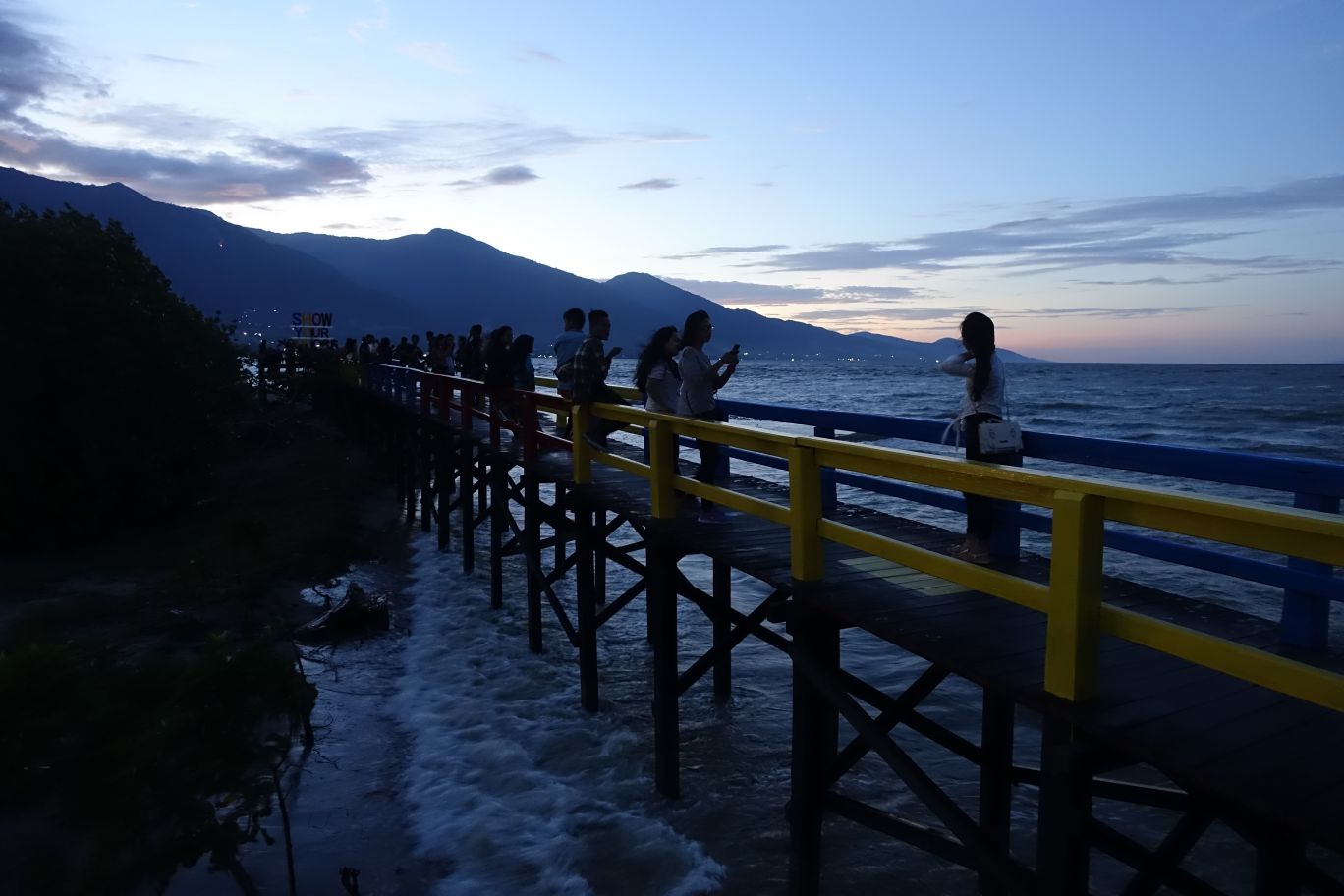 People enjoying the sunset from a colorful pier over the ocean with mountains in the background.