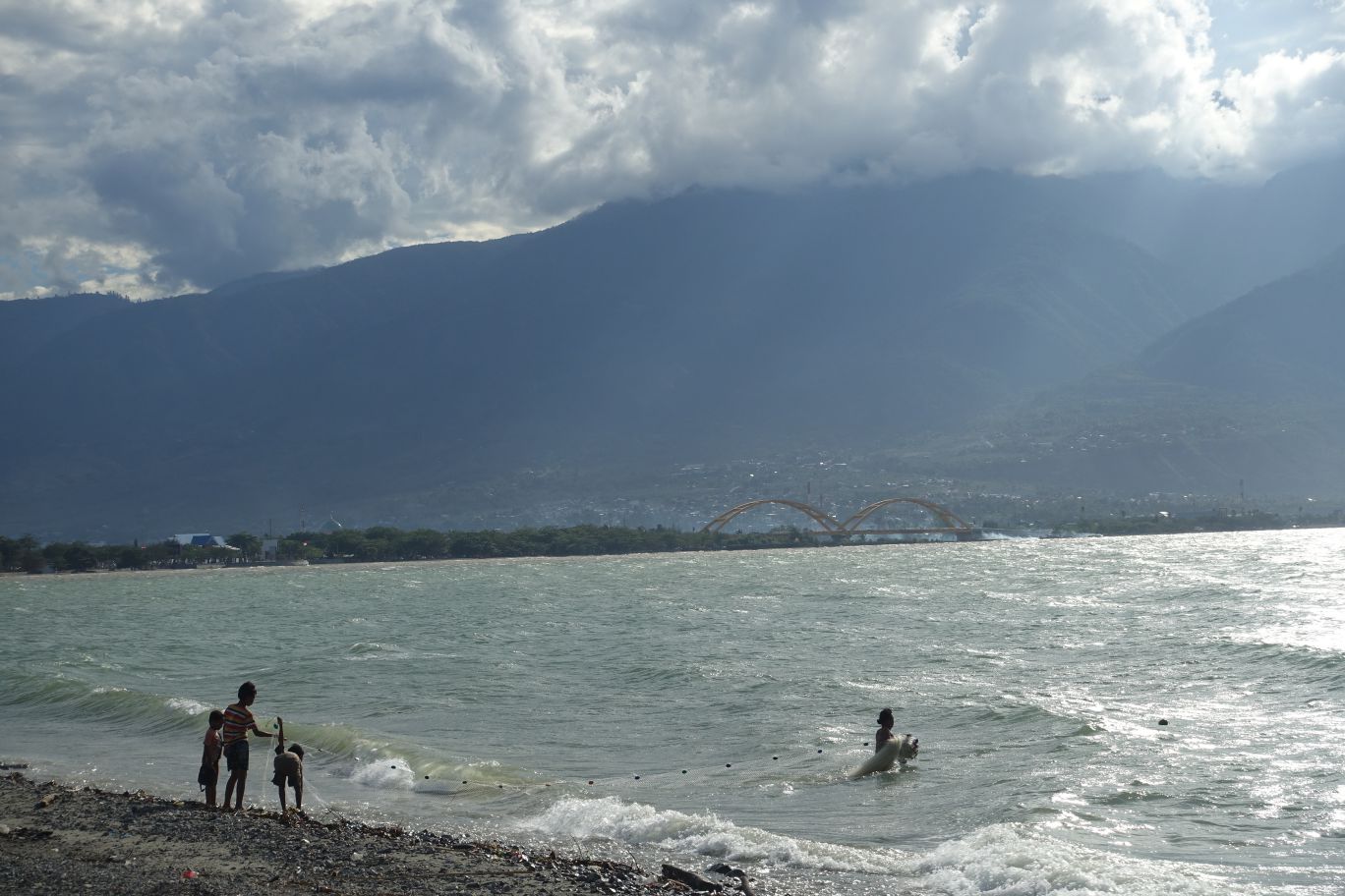 People fishing with a net on a rocky beach, with a city, yellow arched bridge, and mountains under a cloudy sky in the background.