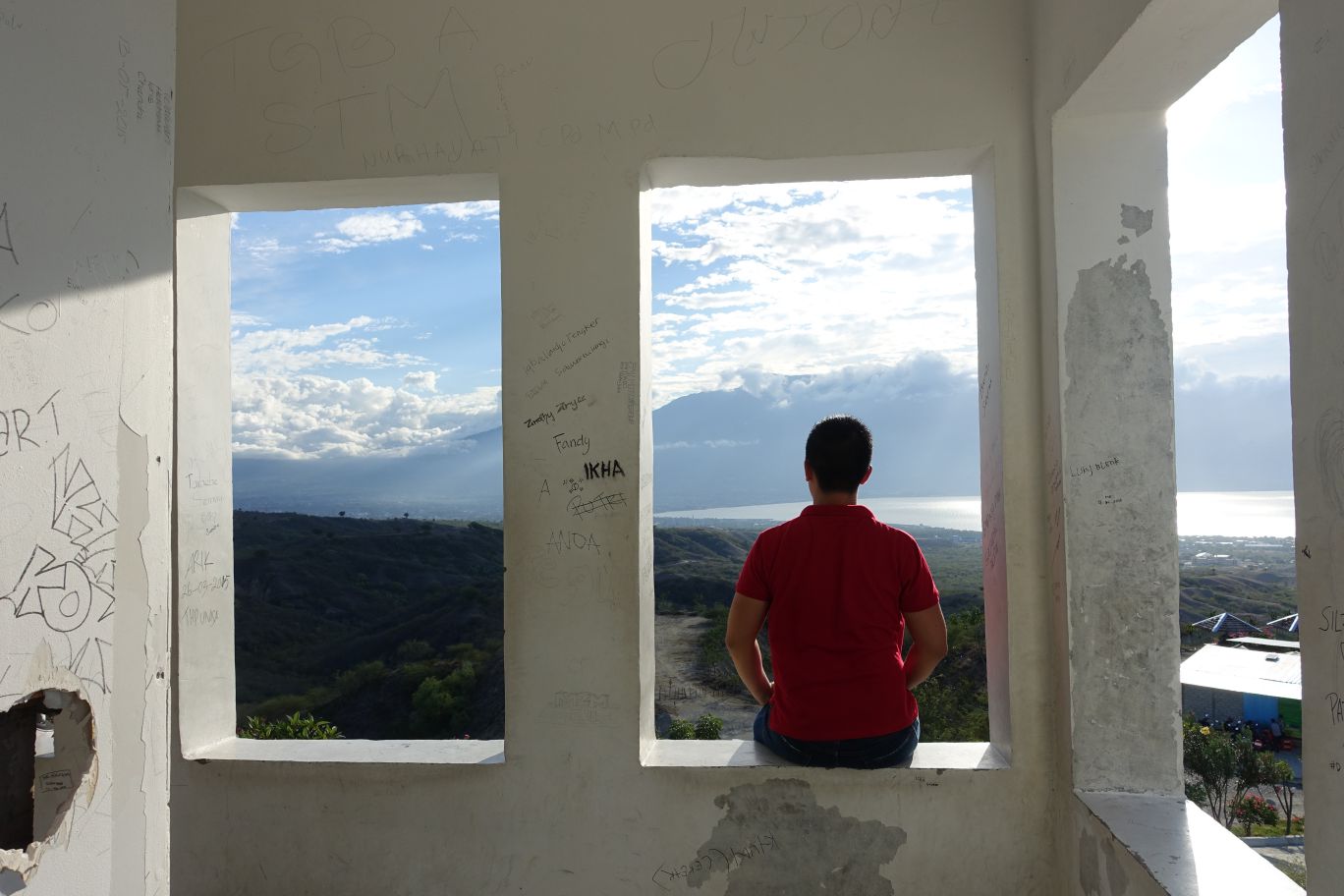 A person in a red shirt sits on a ledge, looking out at a scenic landscape of hills, water, and mountains from a graffiti-covered structure.
