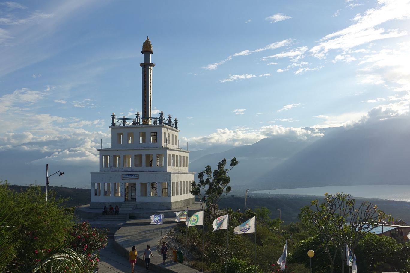 The white tiered Palu City Icon Monument on a hill overlooking Palu Bay and mountains under a partly cloudy sky.