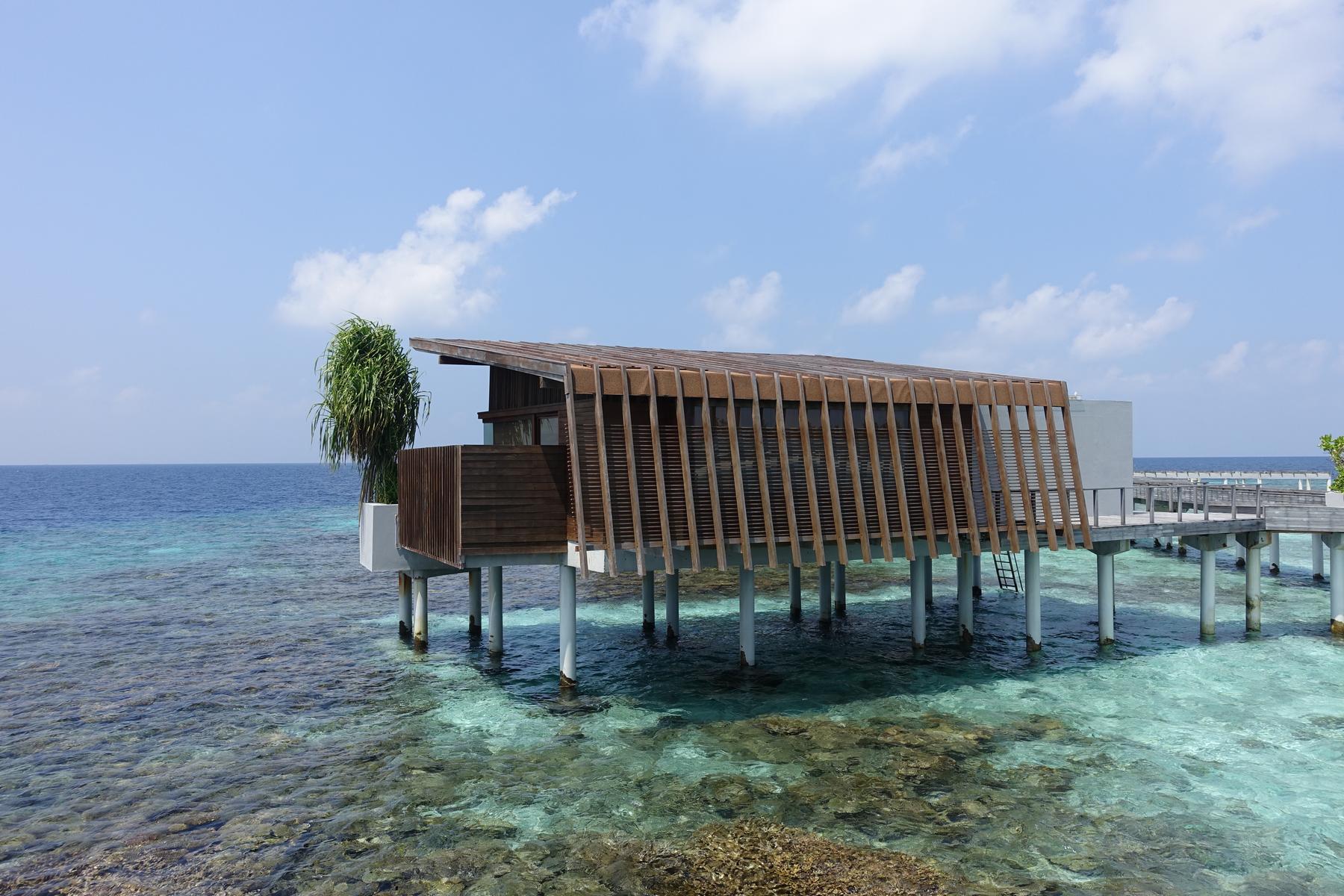 A modern overwater bungalow with a wooden slatted facade overlooks clear turquoise waters in the Maldives.