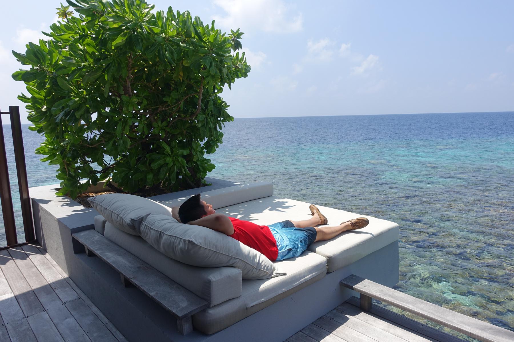 A man relaxes on an outdoor daybed at a water villa overlooking the clear ocean.