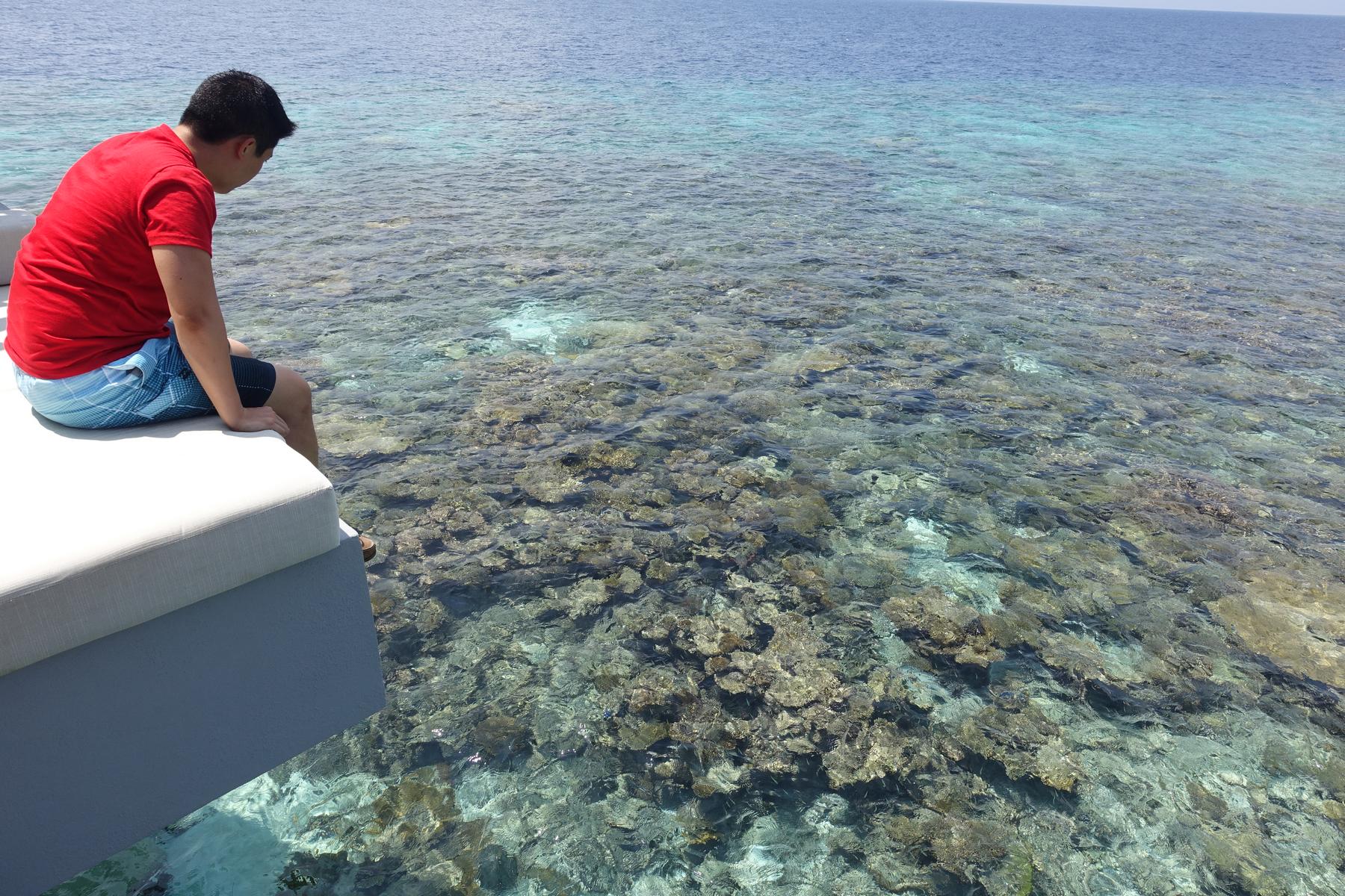 A person sits on the edge of a water villa deck, looking down at the clear turquoise water and coral reef below.