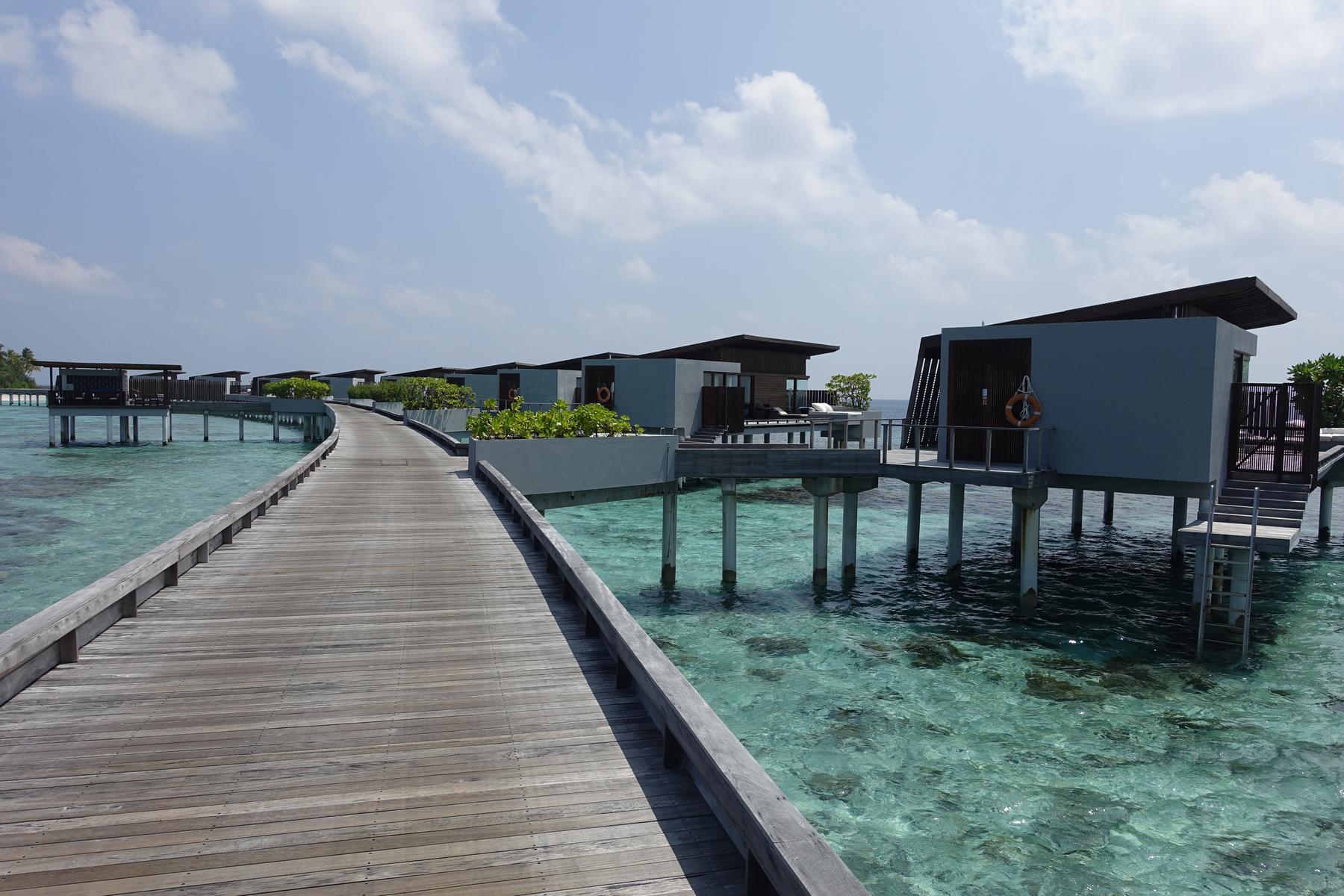 A wooden walkway curves over clear turquoise water, leading to a row of modern overwater bungalows.