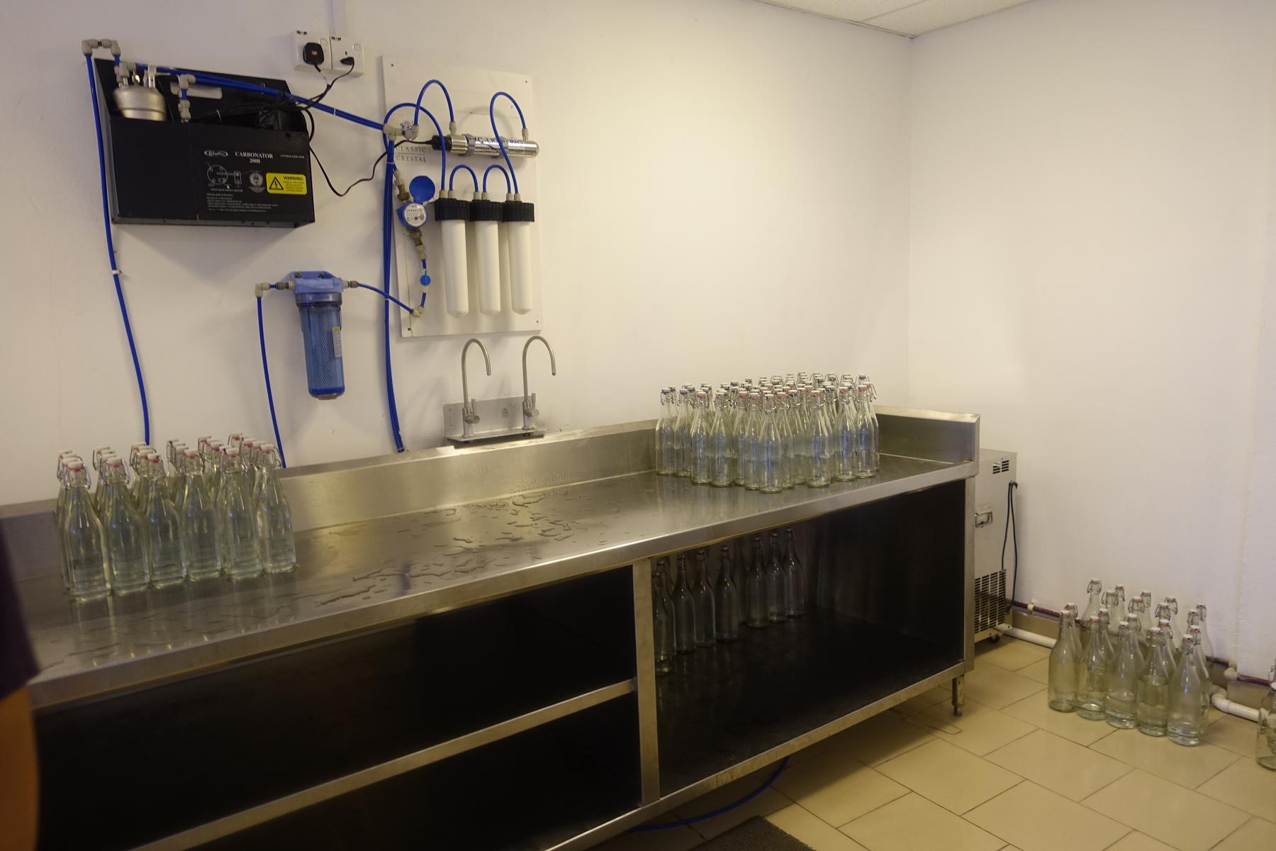 An in-house water bottling station with filtration equipment, taps, and rows of reusable glass bottles on a steel counter.