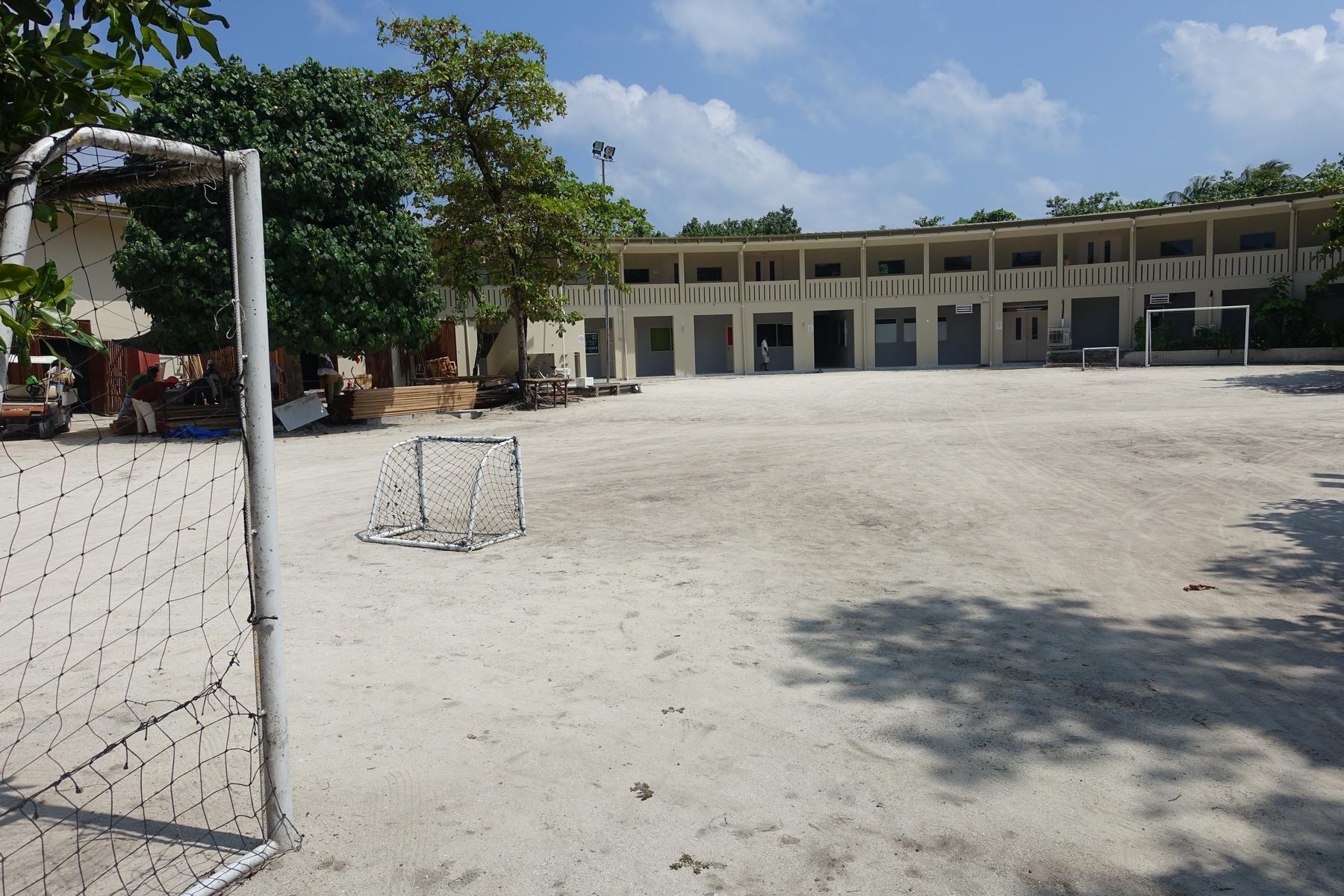 A sandy soccer field with a large goal, a smaller practice goal, and a two-story building in the background.