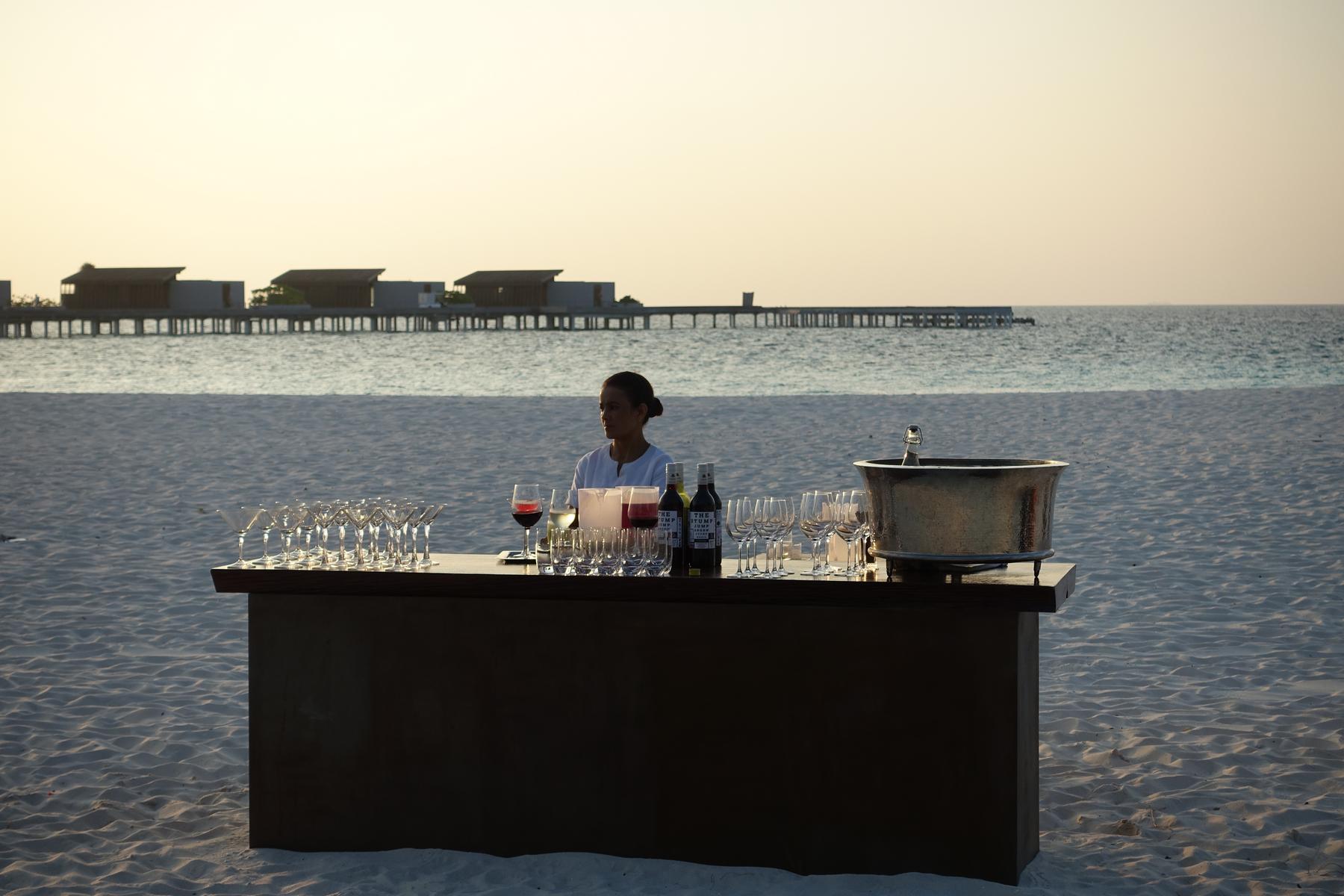 A person stands behind a beach bar with drinks and glasses at sunset in the Maldives.