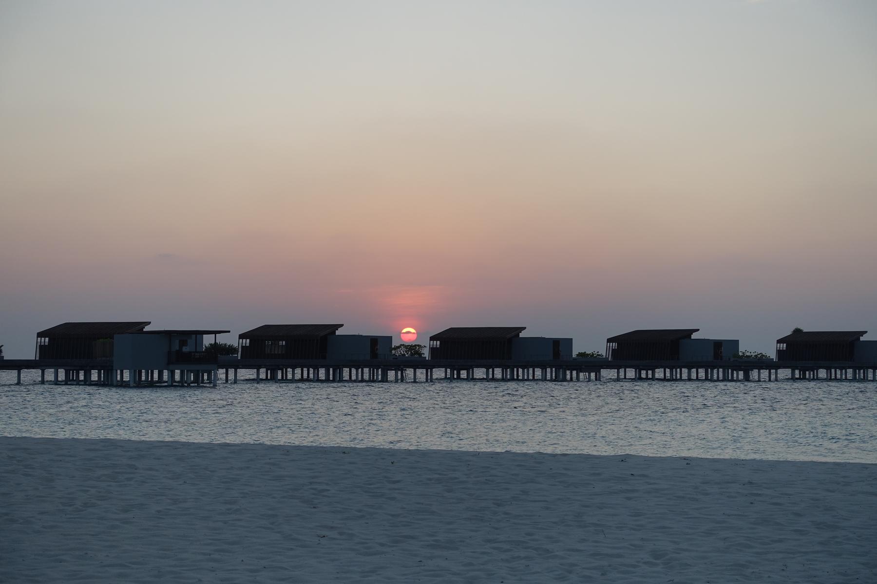 A sandy beach and overwater villas silhouetted against a pink and orange sunset at the Park Hyatt Maldives.