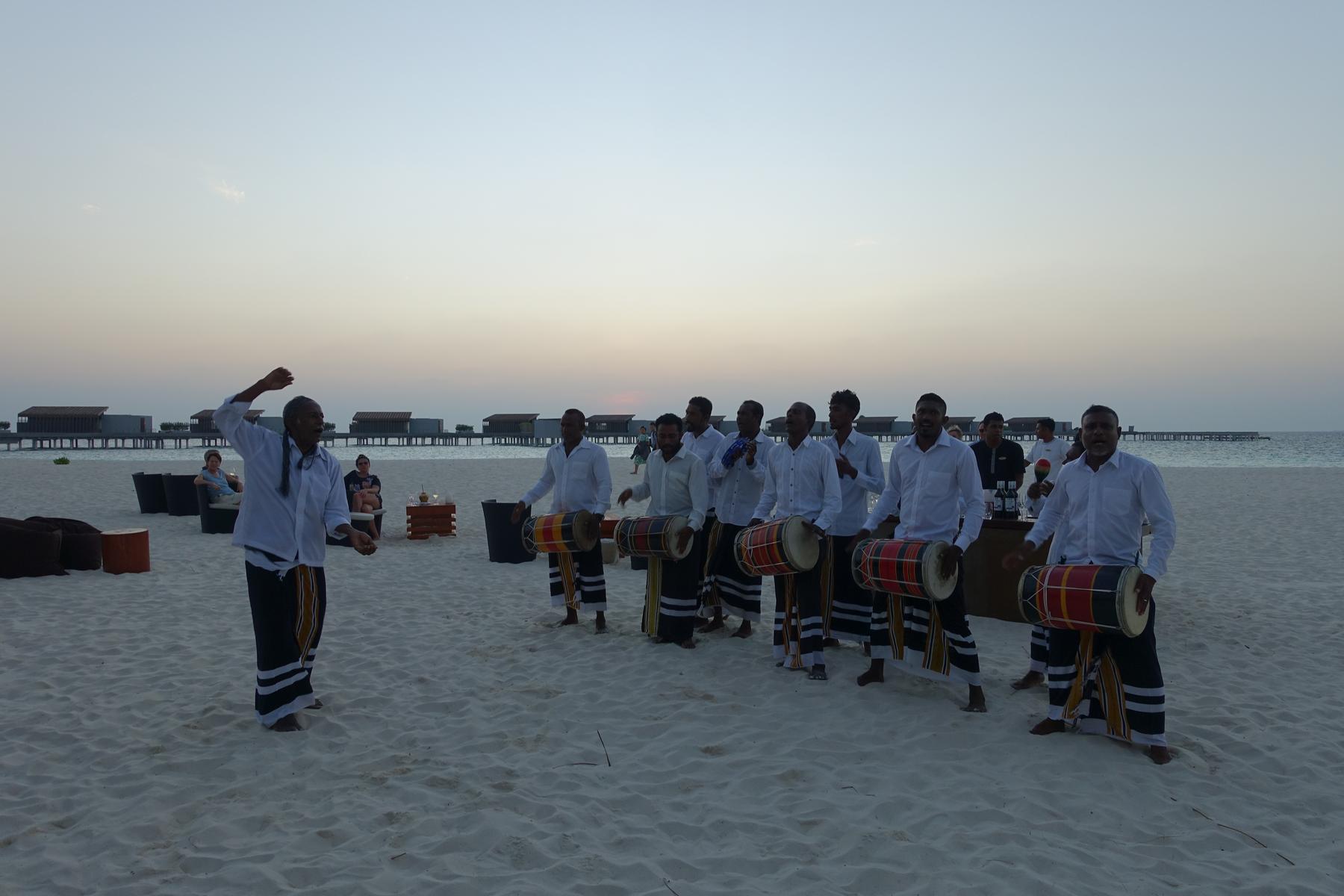 A group of men perform a traditional Bodu Beru drum and dance show on a Maldives beach at dusk, with overwater bungalows in the background.